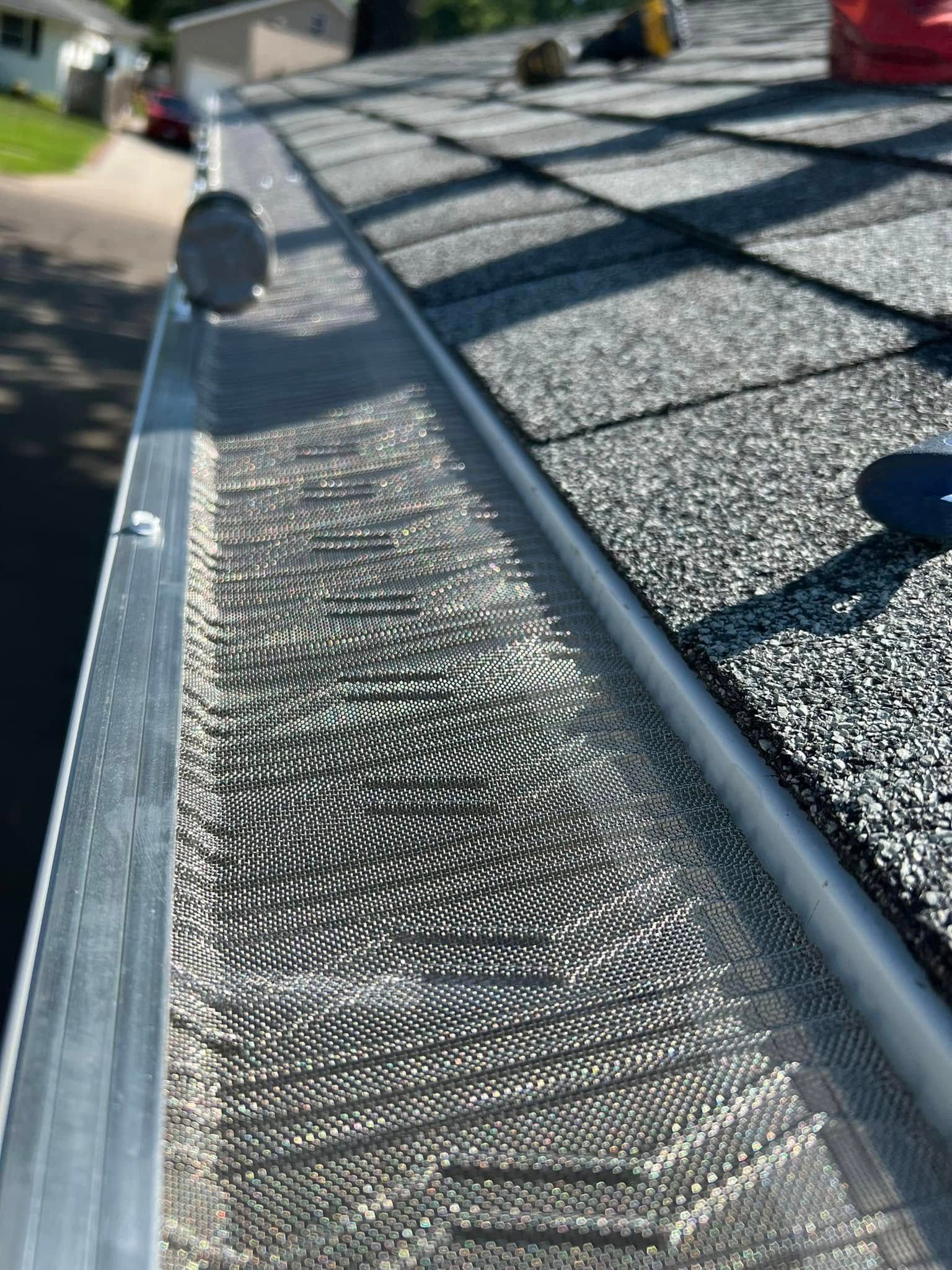 Close-up of a metal rain gutter beside a shingled roof in bright sunlight.