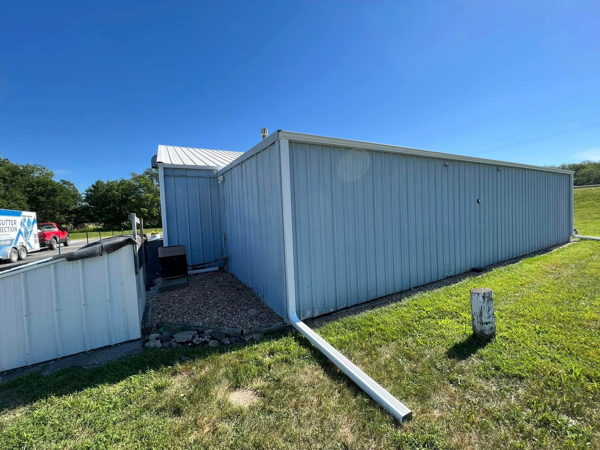 White metal shed with a leaning gutter pipe on a grassy lot under a clear blue sky
