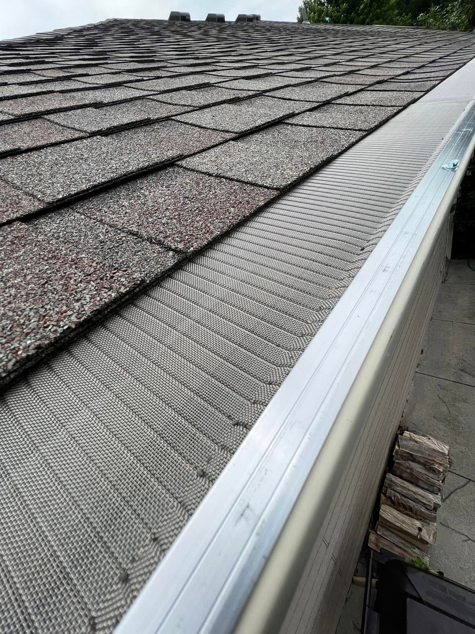 Close-up of a shingled roof edge with a silver gutter and leaf guard mesh.