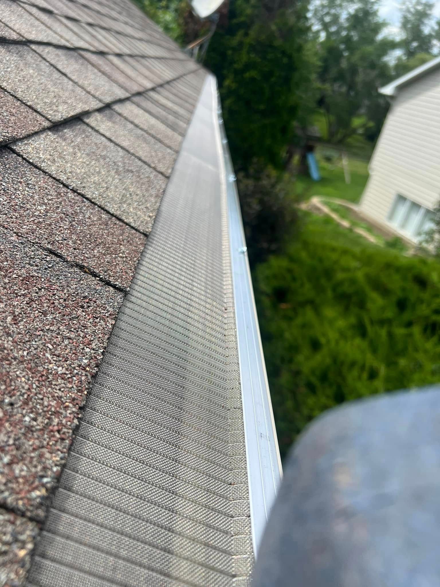 Close-up of roof shingles and a gutter edge beside a house, with trees and yard below.