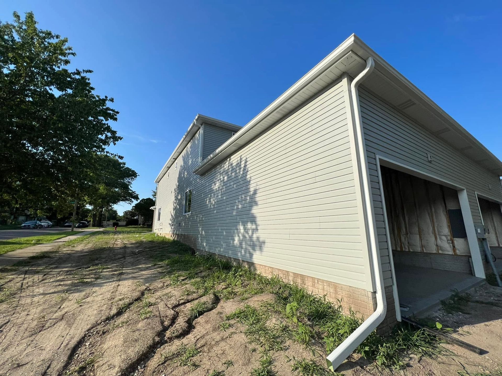 White house exterior with open garage bay, white siding, and a dirt driveway under a blue sky.