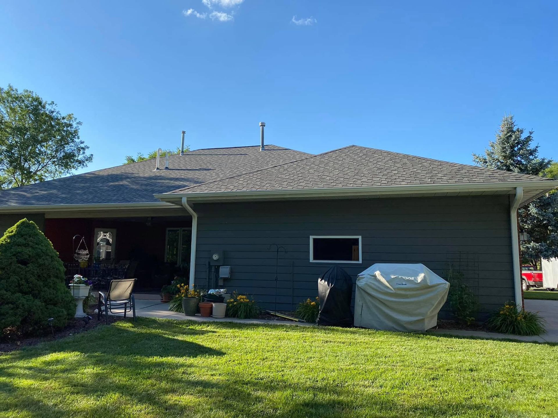 Single-story gray house with a covered patio, green lawn, and covered grill under a blue sky