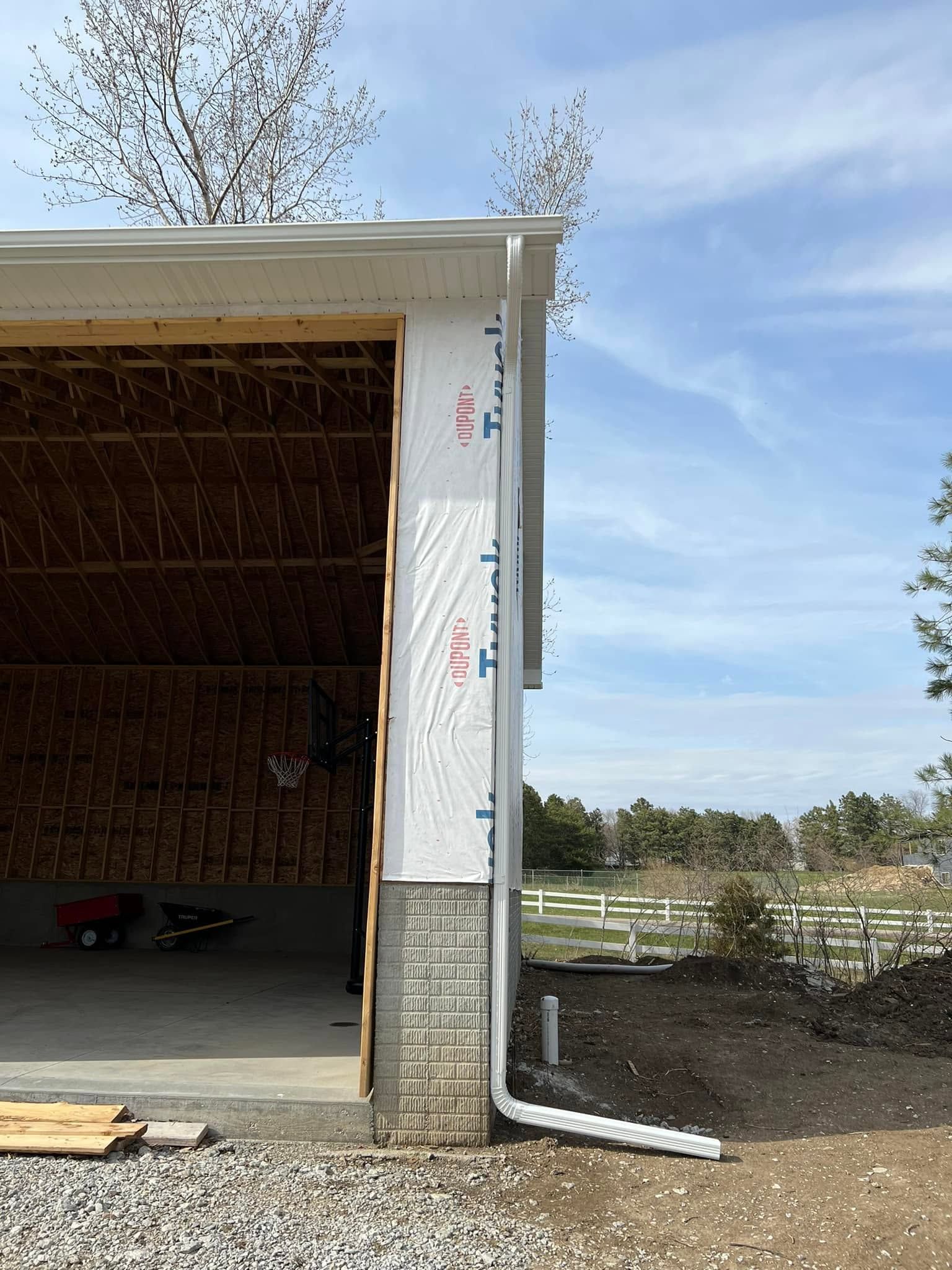 Open garage framed in white siding with plywood interior and a downspout beside a muddy field.
