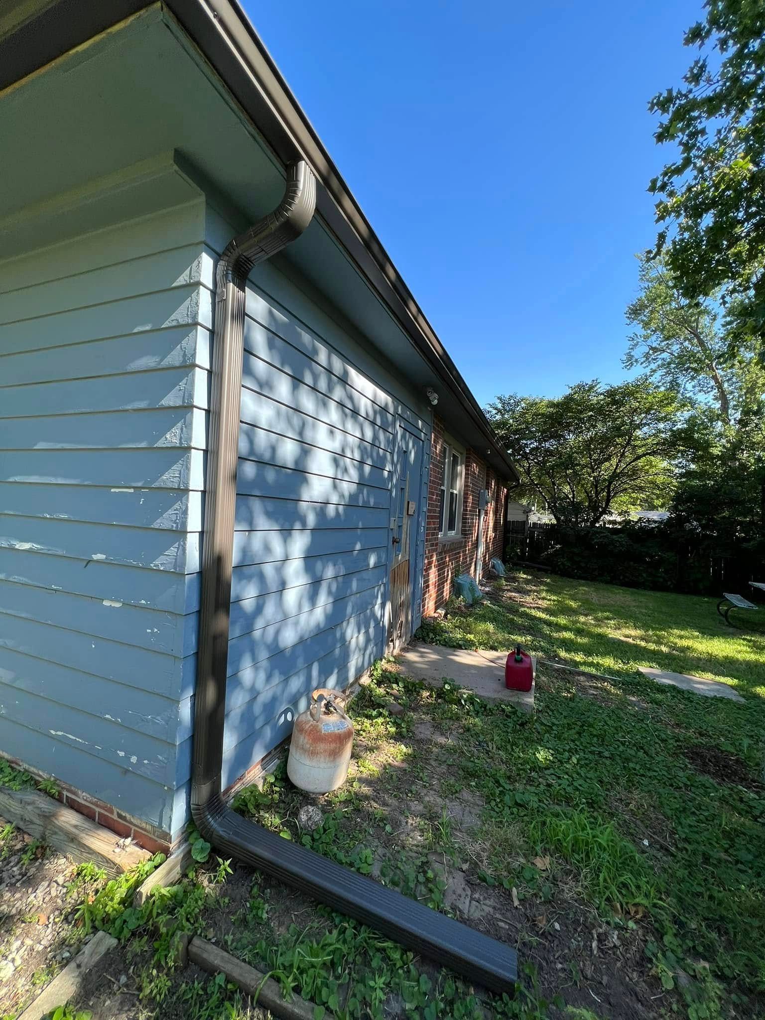 Side of a blue house with white trim, a yard, and potted plants along the wall in bright sunlight.