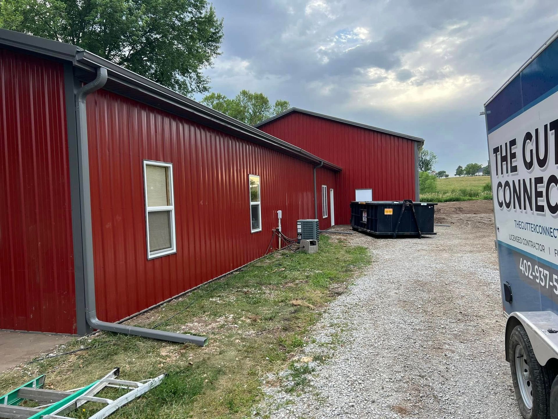 Red metal building beside gravel driveway, with a white trailer and cloudy sky.
