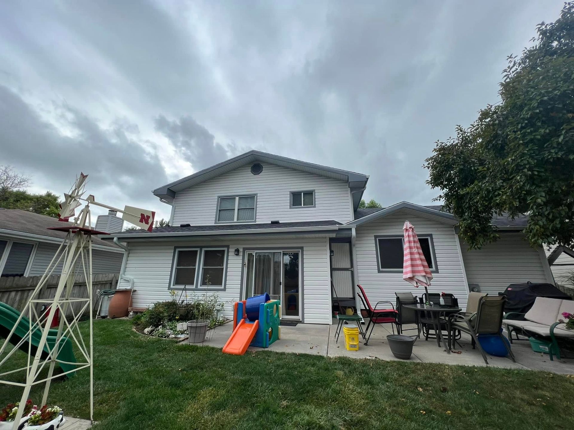 Backyard patio with a two-story house, lawn, patio furniture, umbrella, and children’s slide under a cloudy sky