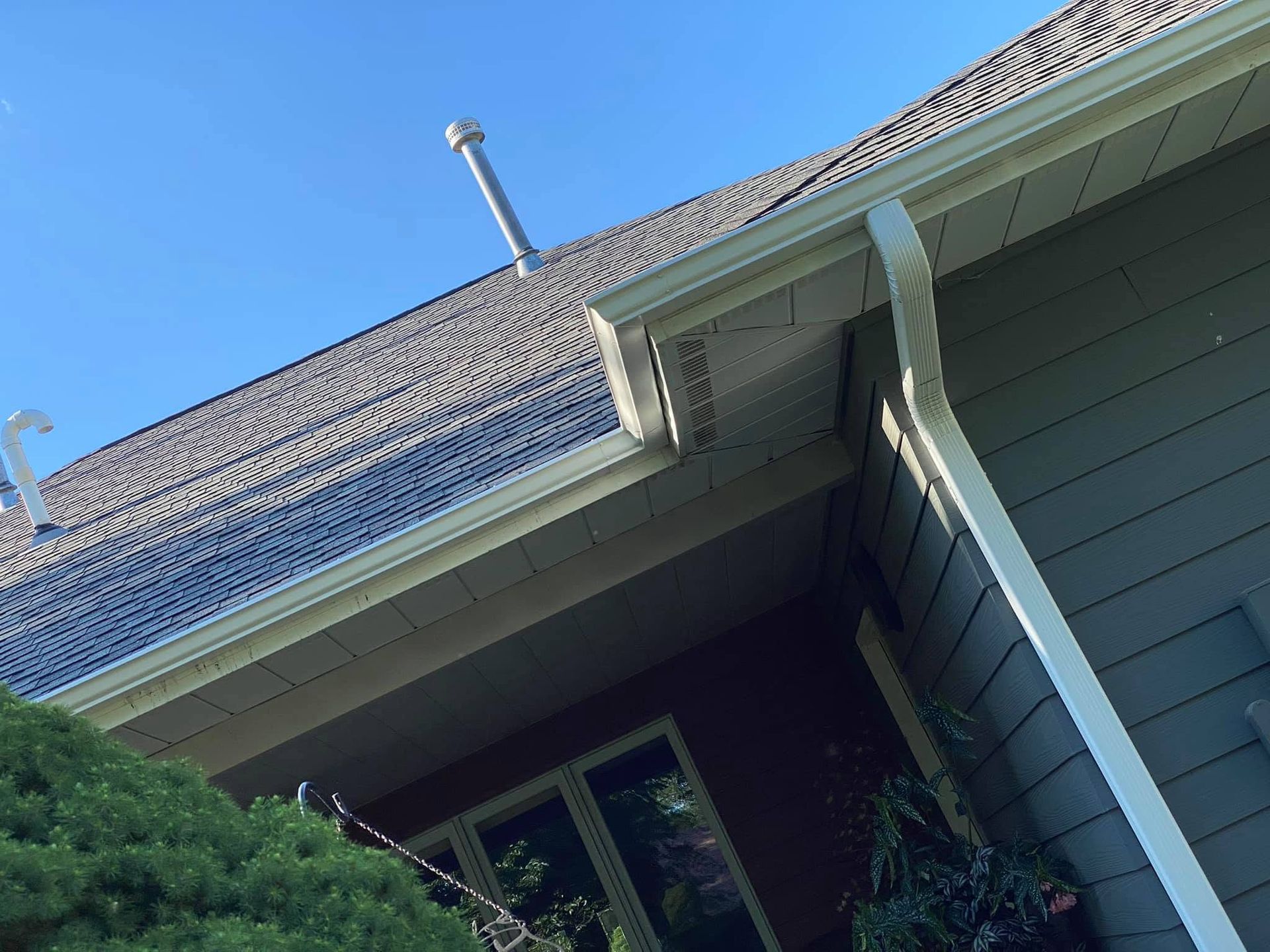 Tilted view of a house roof and gutter against a blue sky, with green siding and trees below