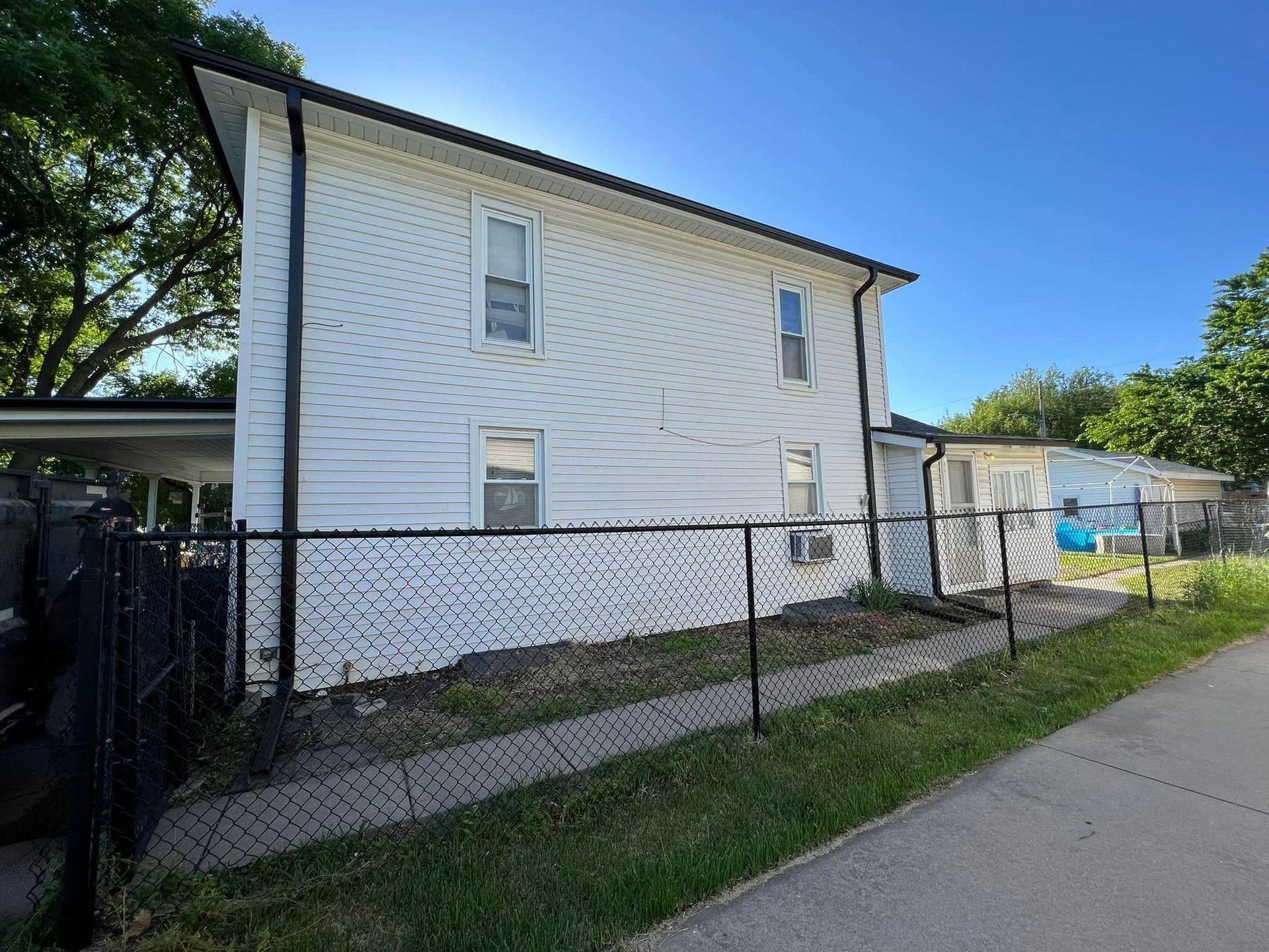 White two-story house with chain-link fence beside a sidewalk under a clear blue sky
