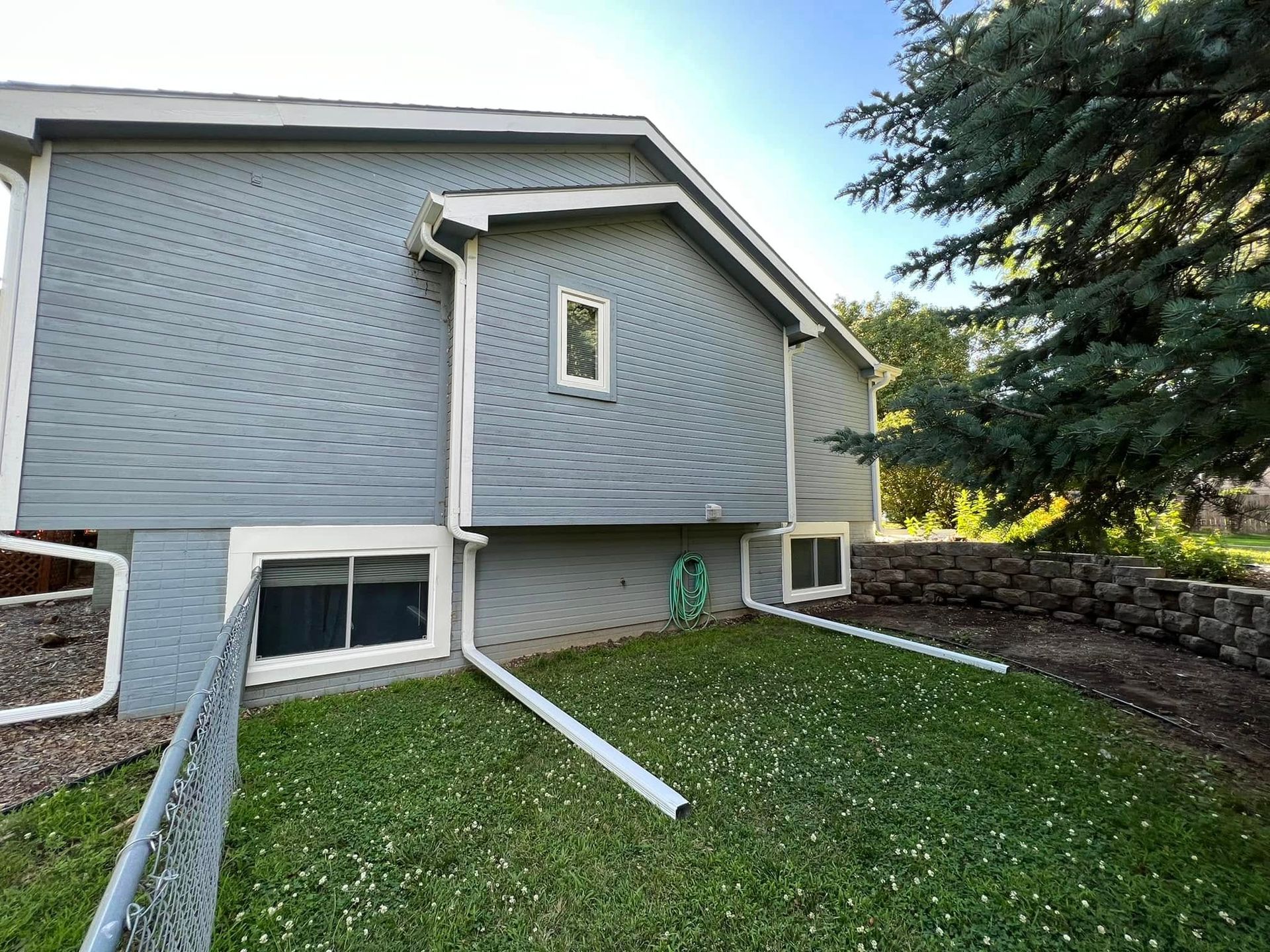 Blue house exterior with basement windows, sloped lawn, and a large evergreen tree on the right