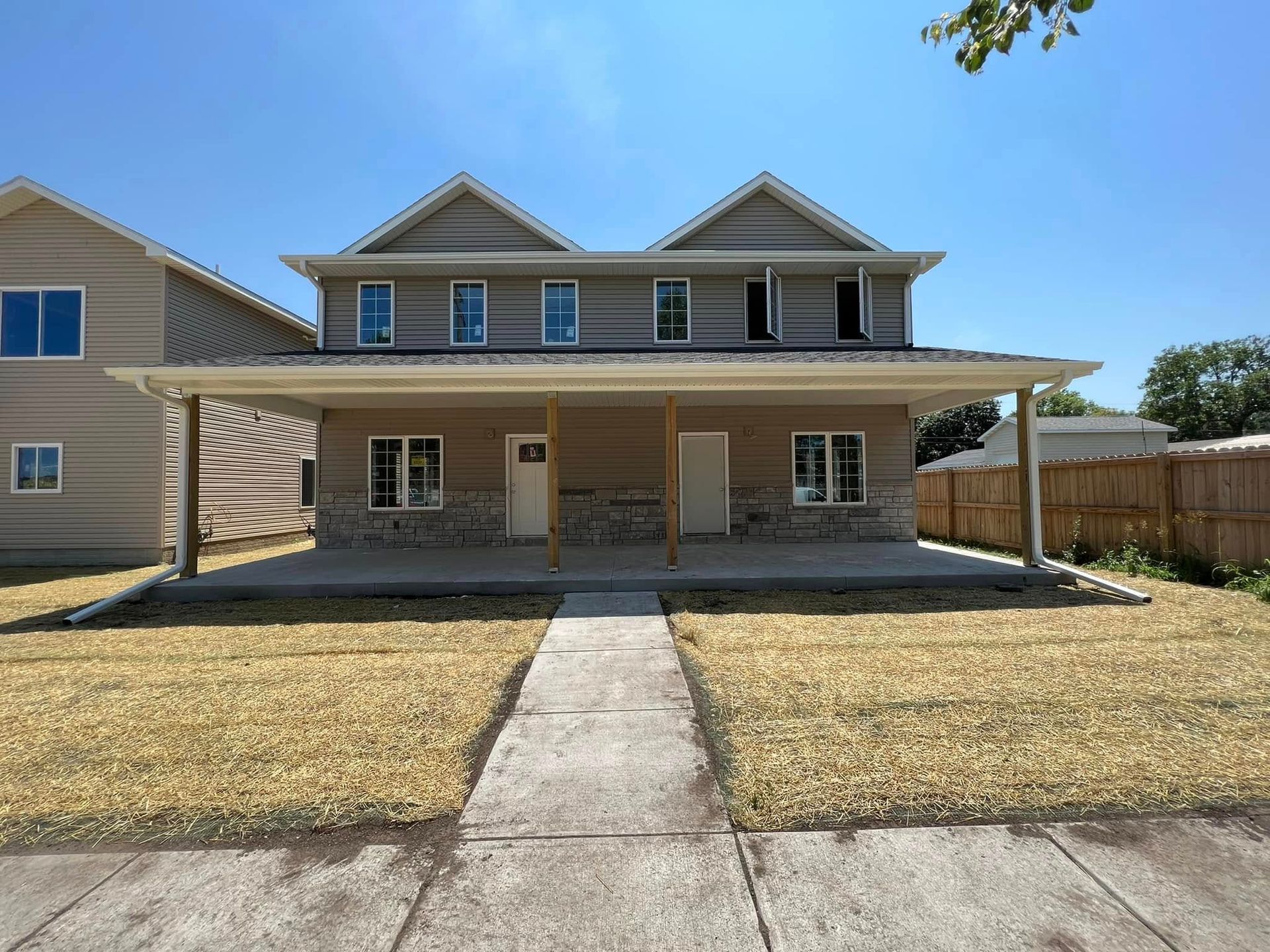Two-story beige house with a covered front porch, centered walkway, and dry lawn under a clear blue sky