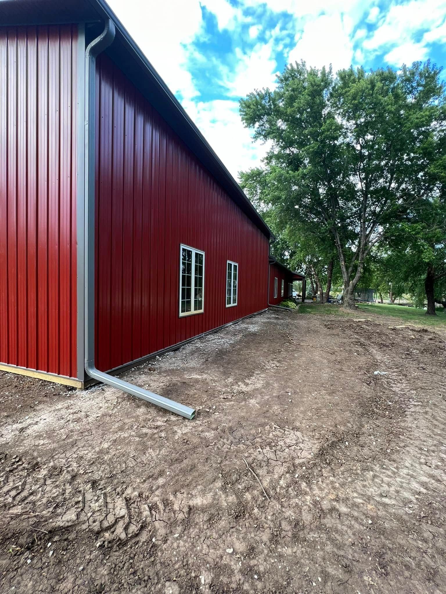 Red metal barn with white-framed windows beside a dirt path and trees under a cloudy sky