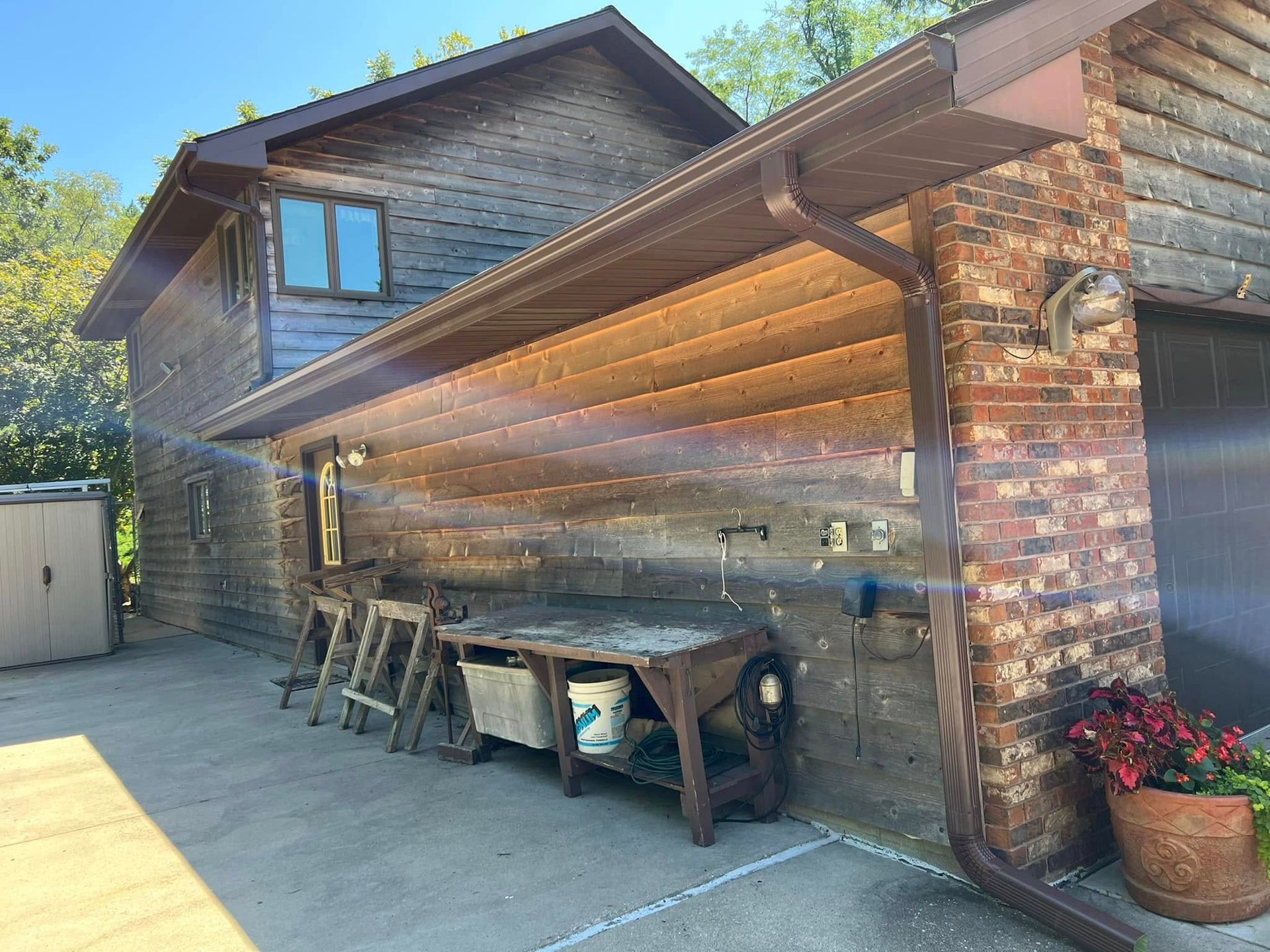 Covered outdoor patio beside a brick house, with a wooden picnic table, chairs, and potted flowers.