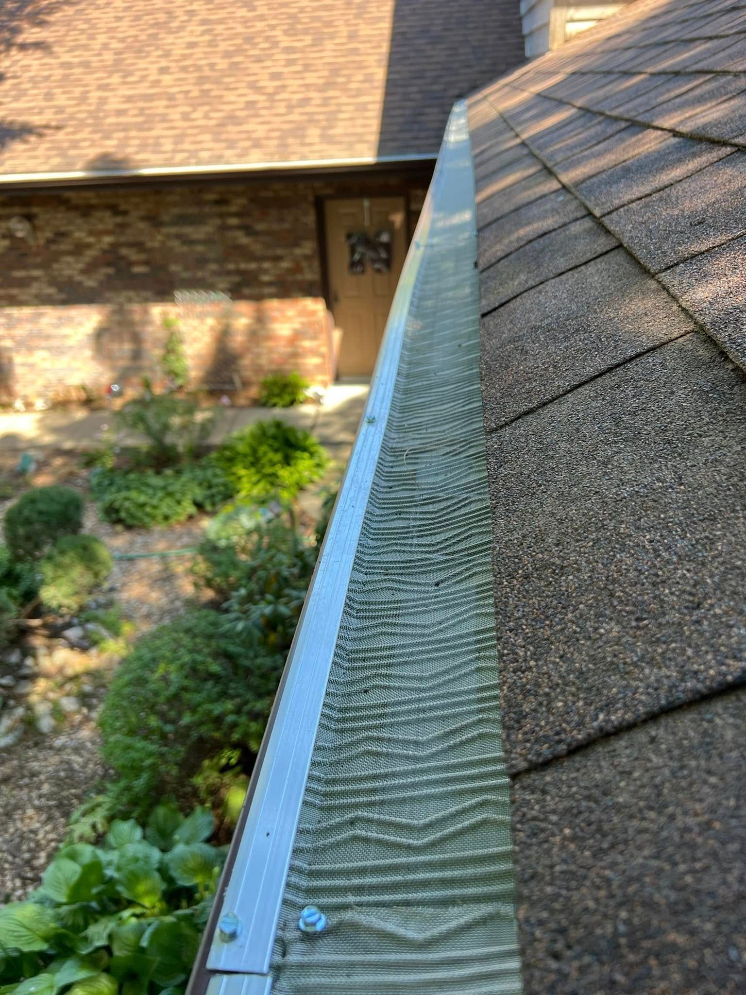Gutter with leaf guards along a shingled roof beside a brick house and garden.