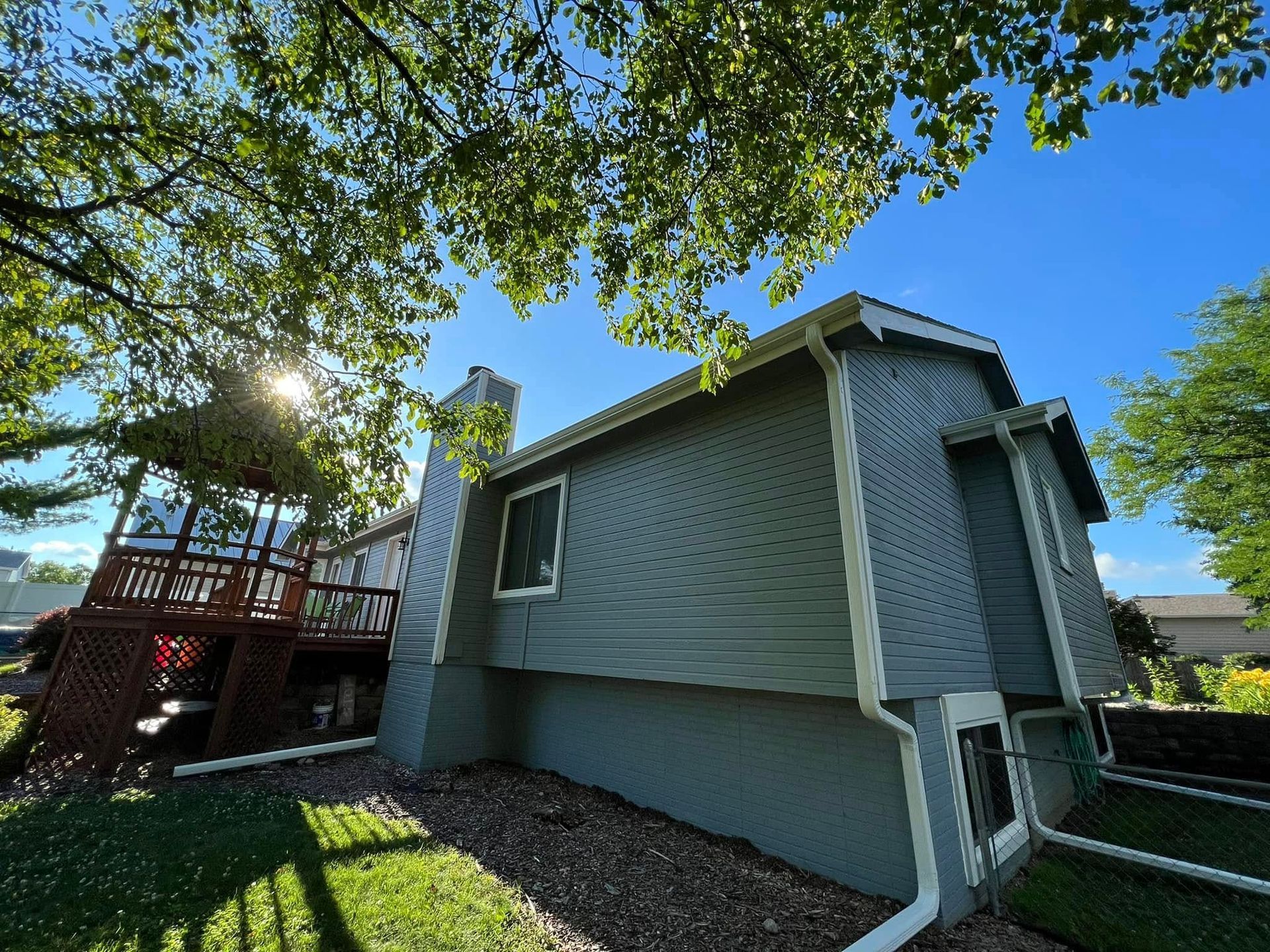 Gray house exterior with deck, trees overhead, and blue sky