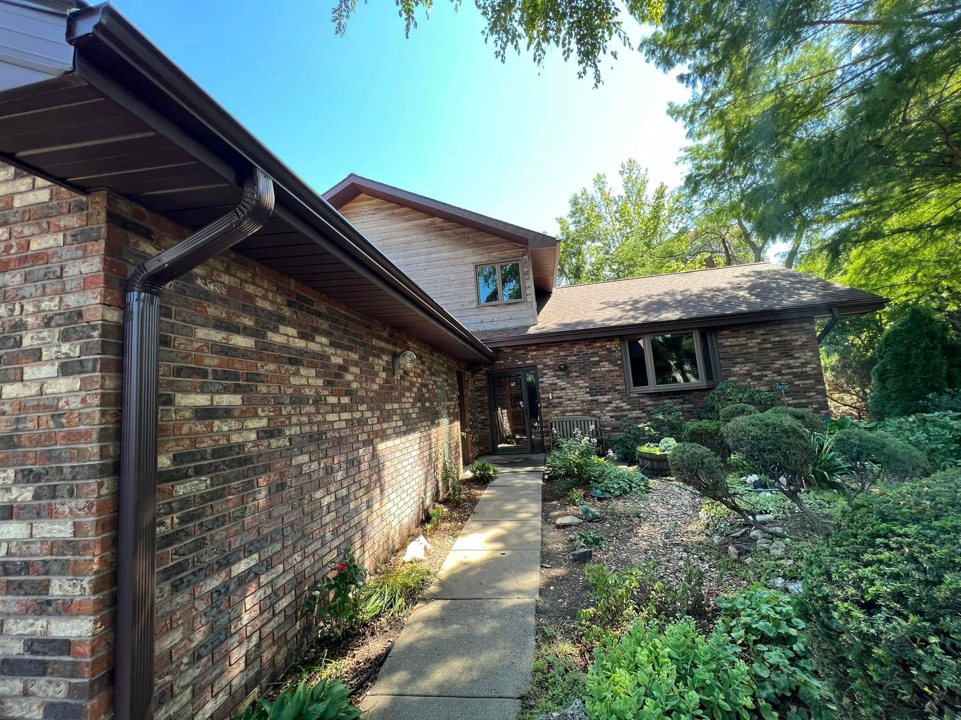 Brick house exterior with a narrow walkway, overhanging roof, and lush green garden plants.