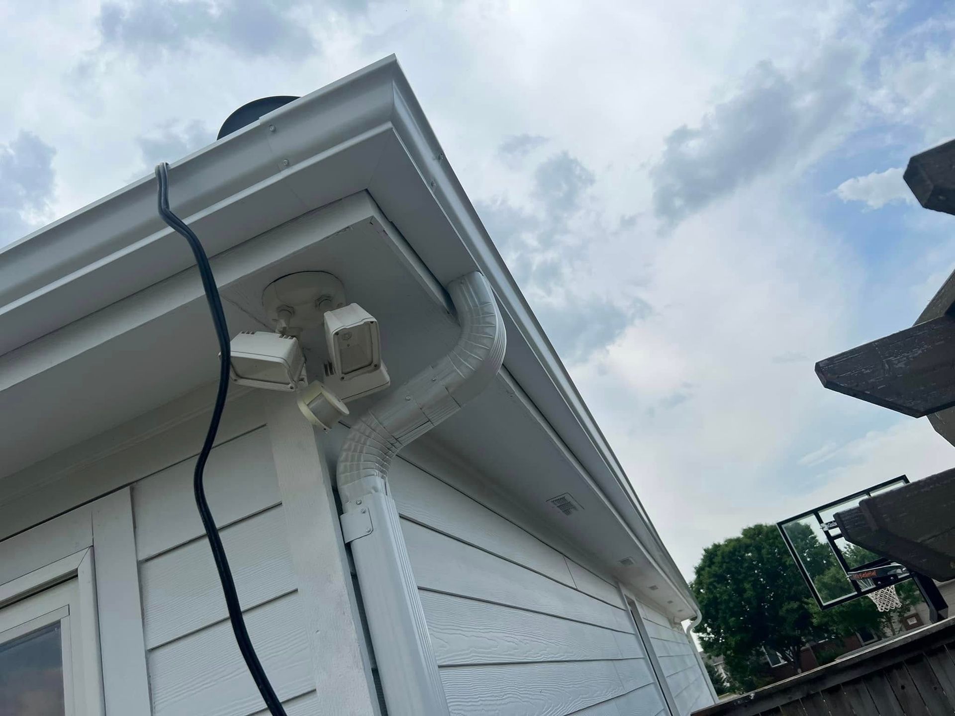 White house eave with mounted security camera and hanging black cable under cloudy sky