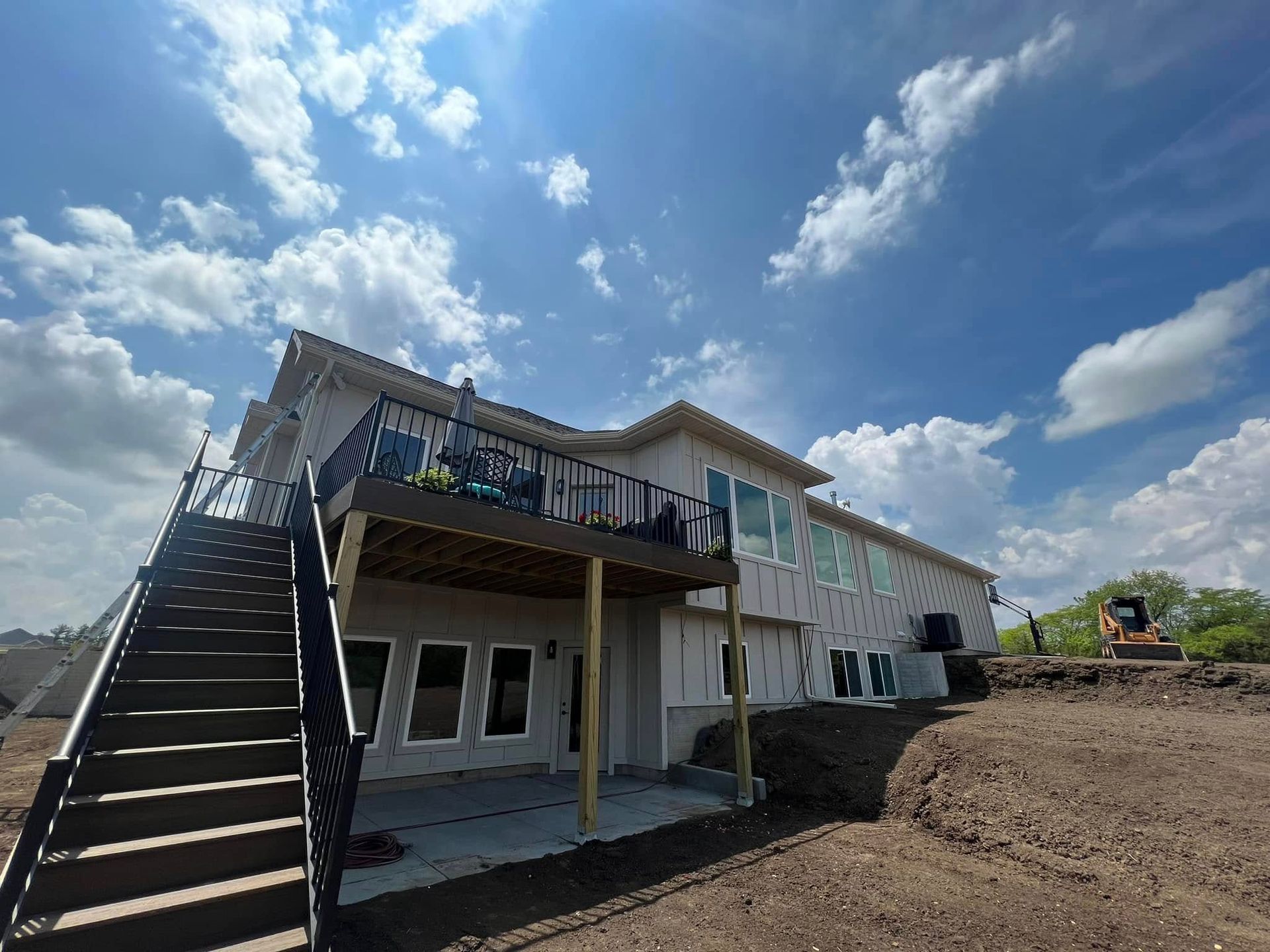 Two-story house under construction with exterior stairs and a balcony, on a dirt lot under a blue sky