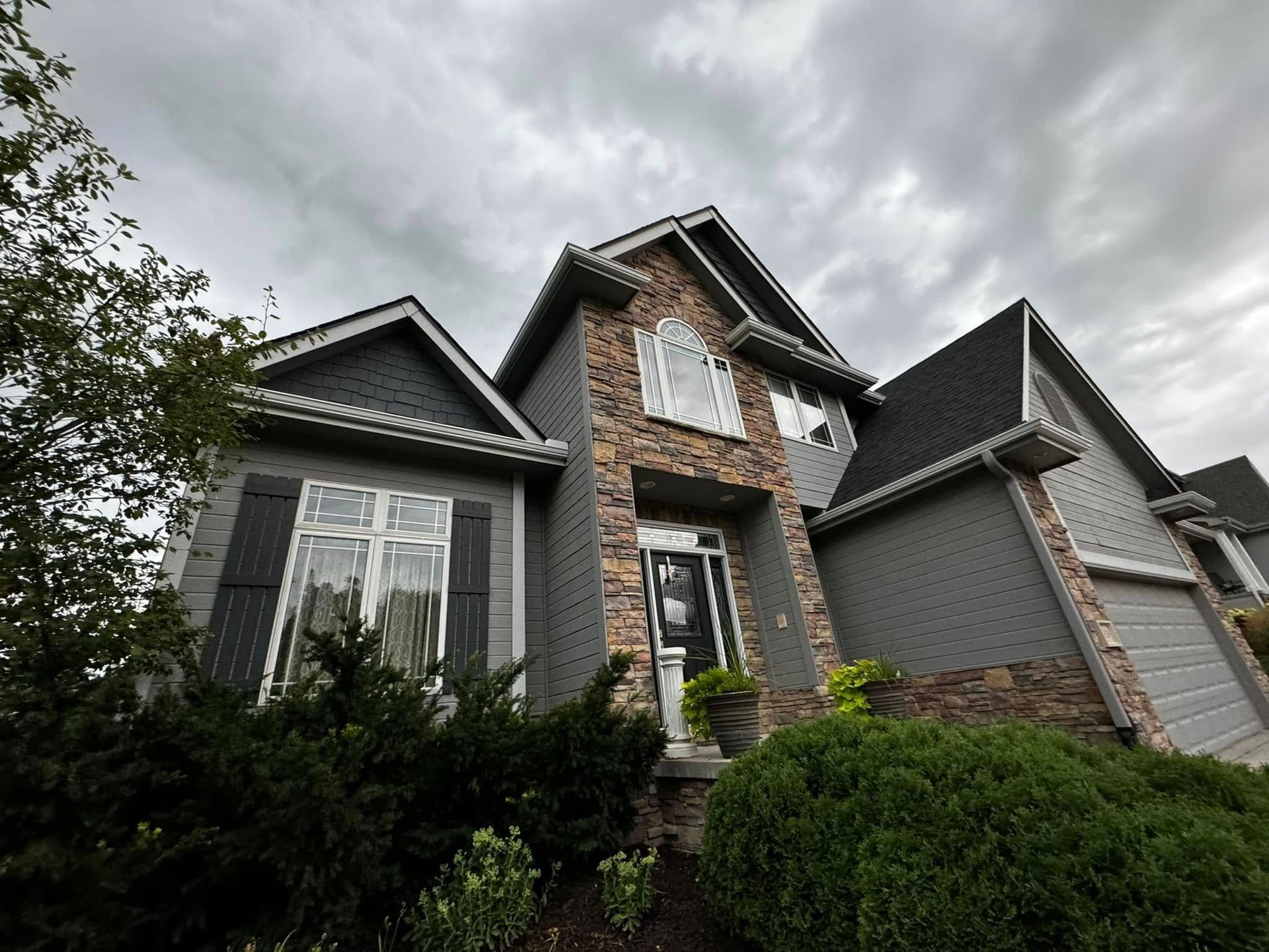 Gray suburban house with stone accents and a cloudy sky, viewed from the front yard