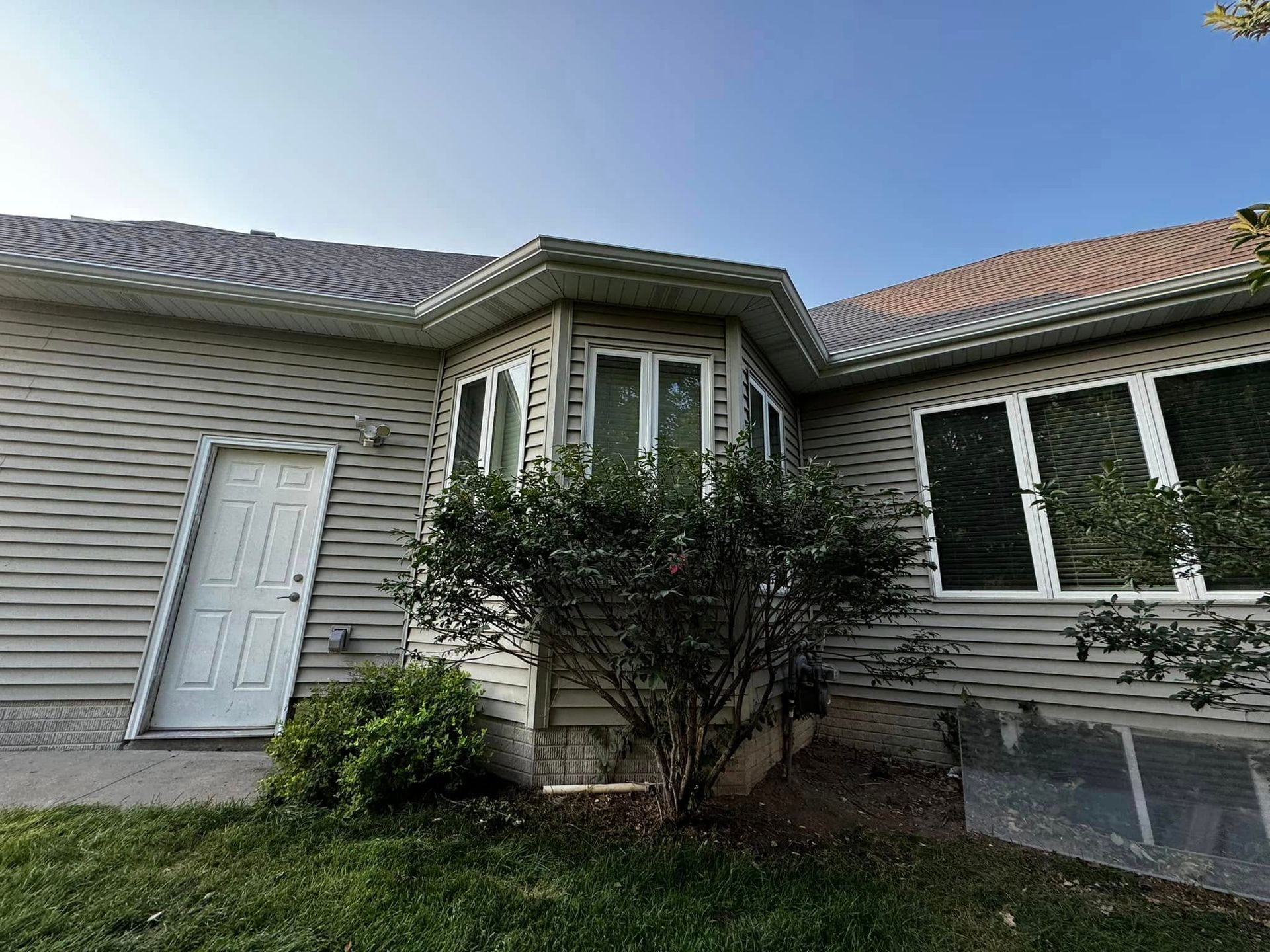 Front entrance of a beige house with a white door, bay window, shrubs, and a tiled roof.