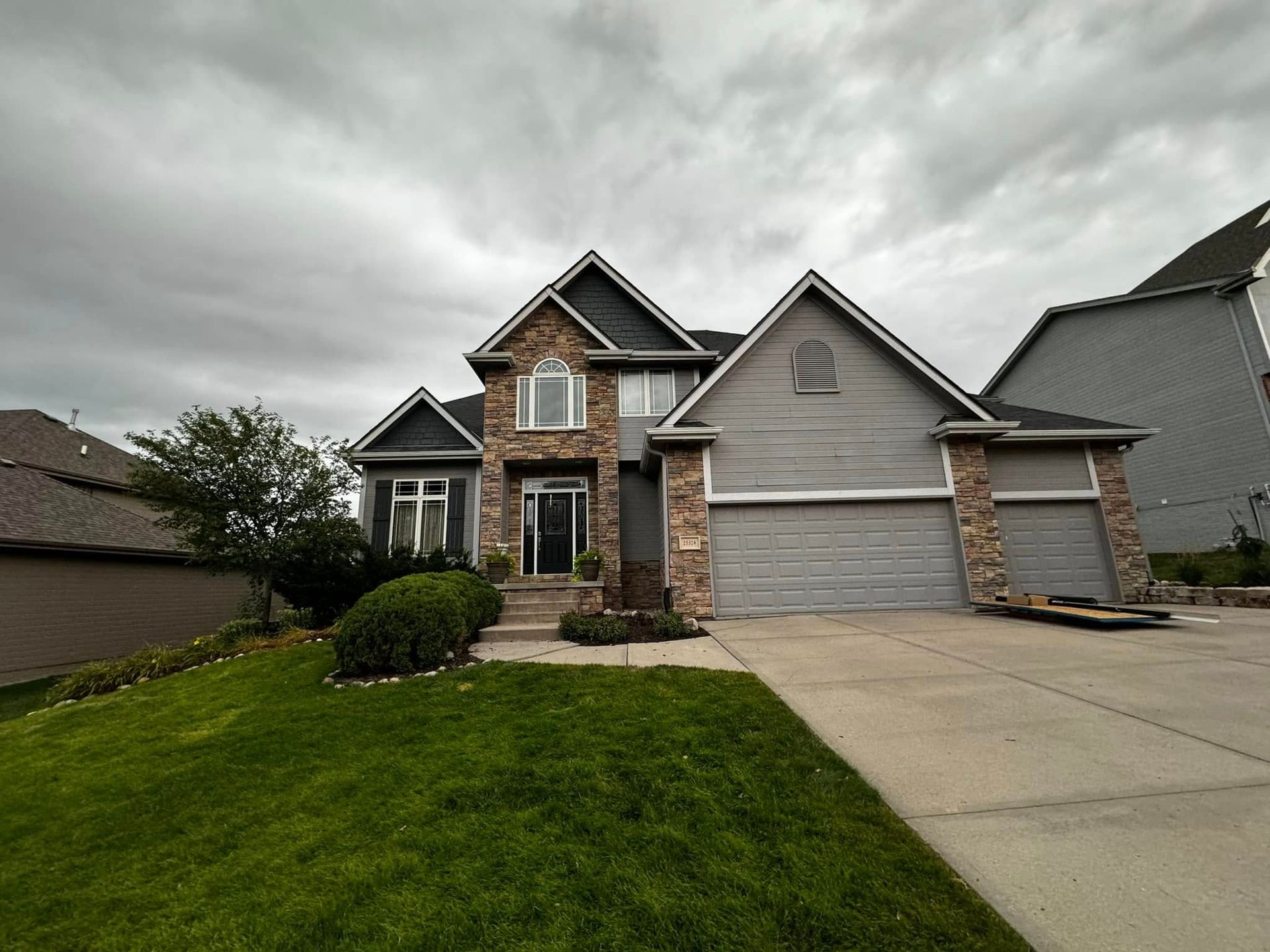 Suburban stone-and-siding house with a two-car garage, front lawn, and concrete driveway under cloudy skies