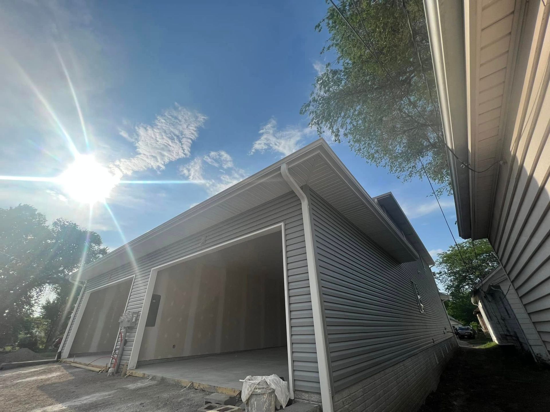 Open garage with white siding under bright sun and blue sky, with trees and a white dog near the entrance