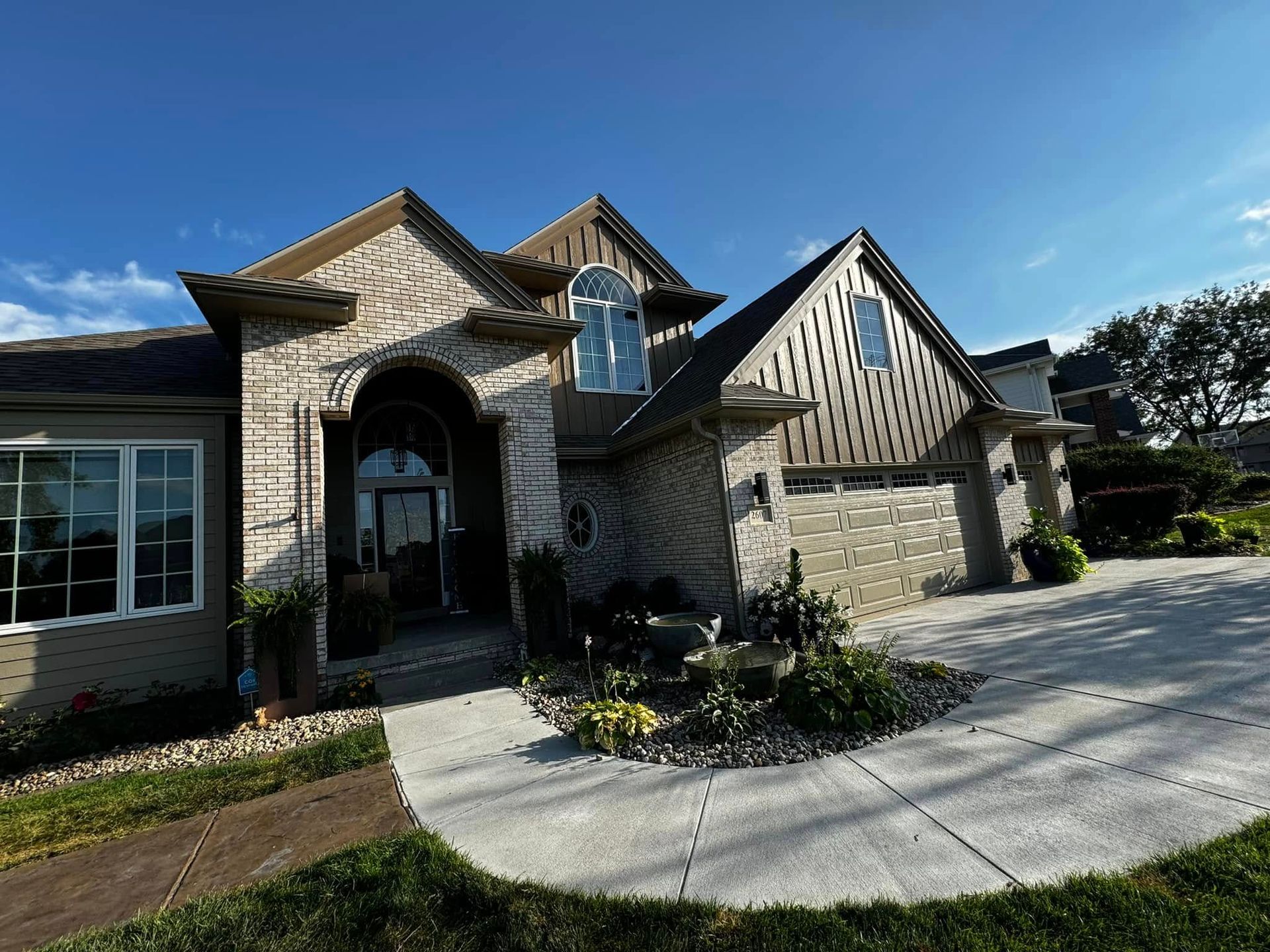 Stone suburban house with arched entry, large windows, and a curved concrete driveway under a blue sky