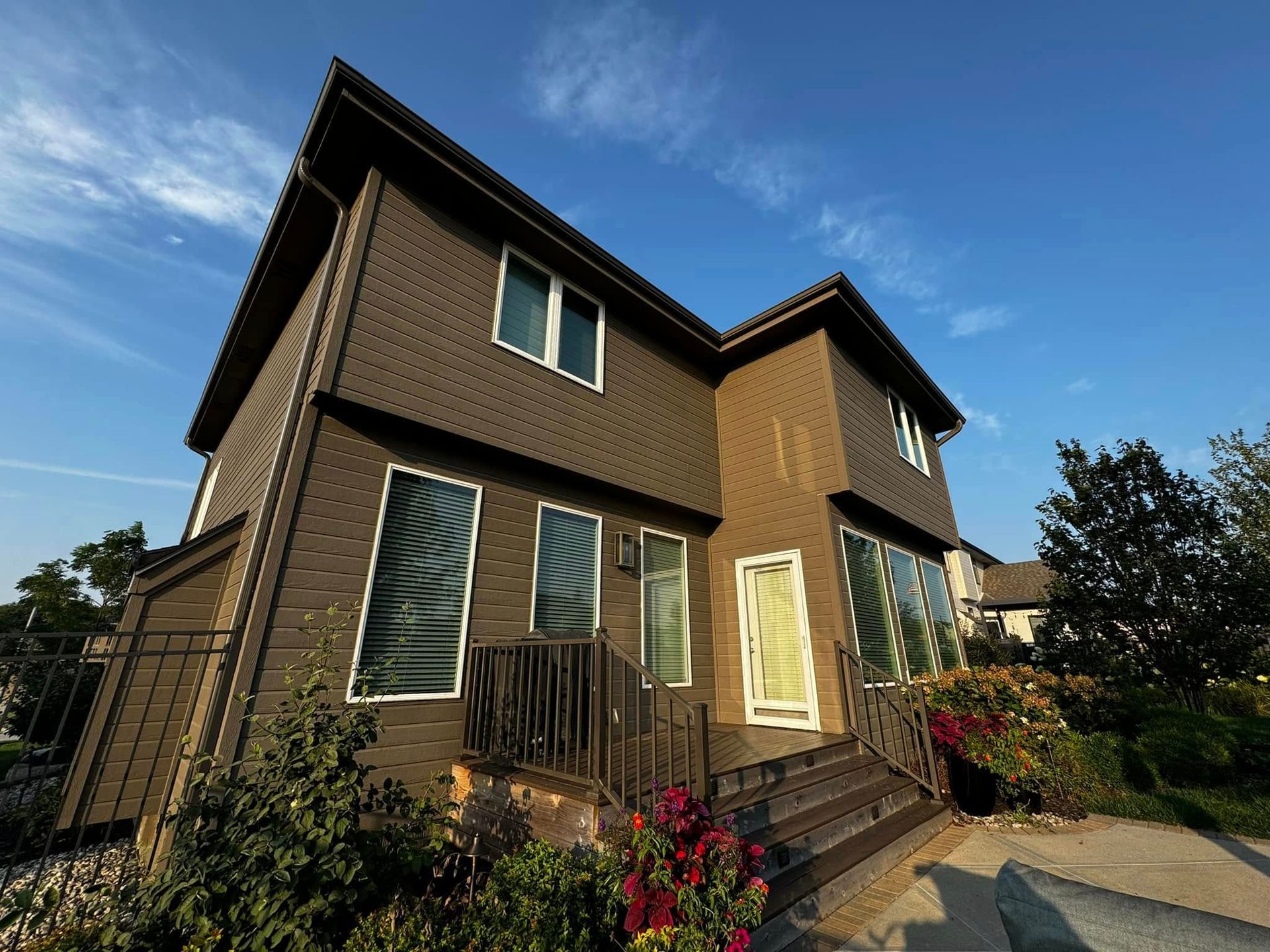 Two-story brown house with large windows, front steps, and a garden under a blue sky.