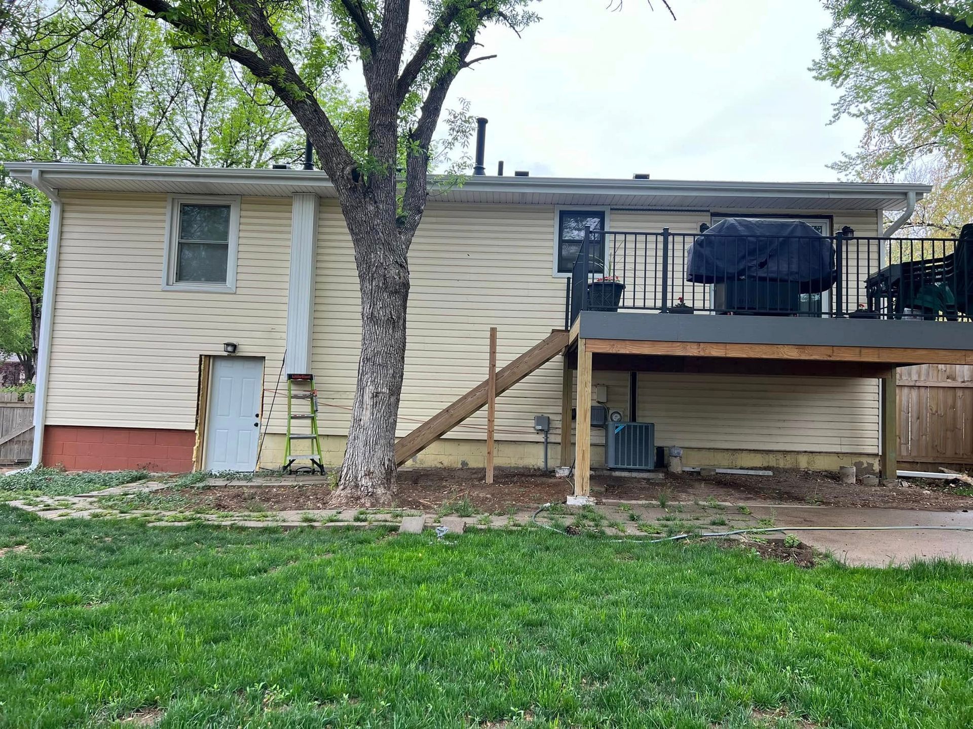 Single-story beige house with a raised deck, staircase, and large tree in the front yard