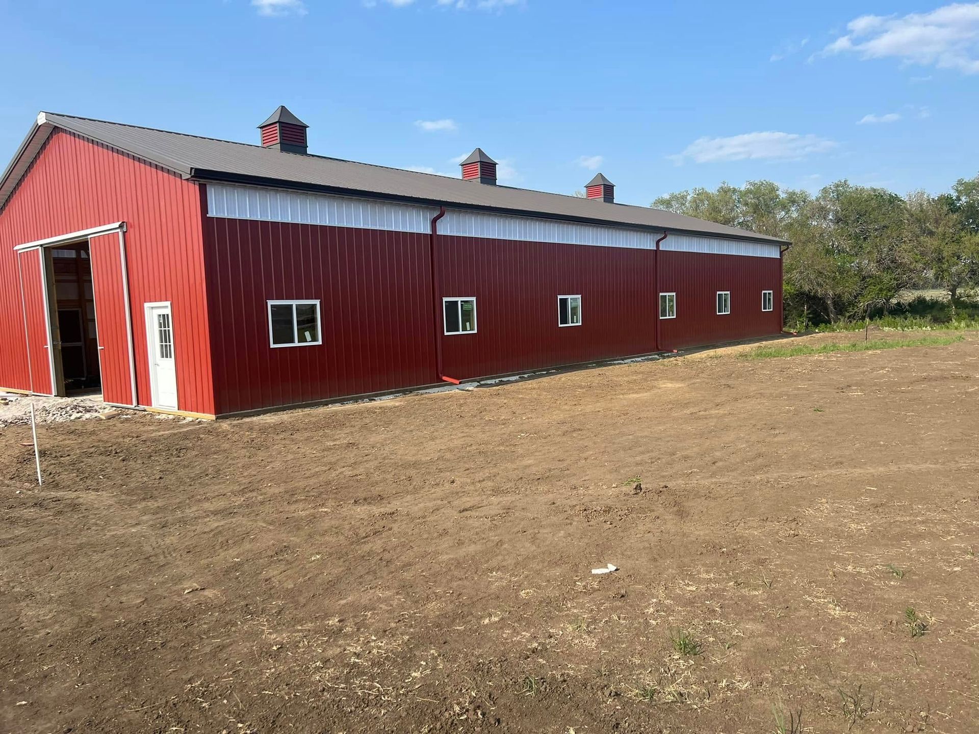 Red barn-style building with a gray roof and small windows on a bare dirt lot under a blue sky