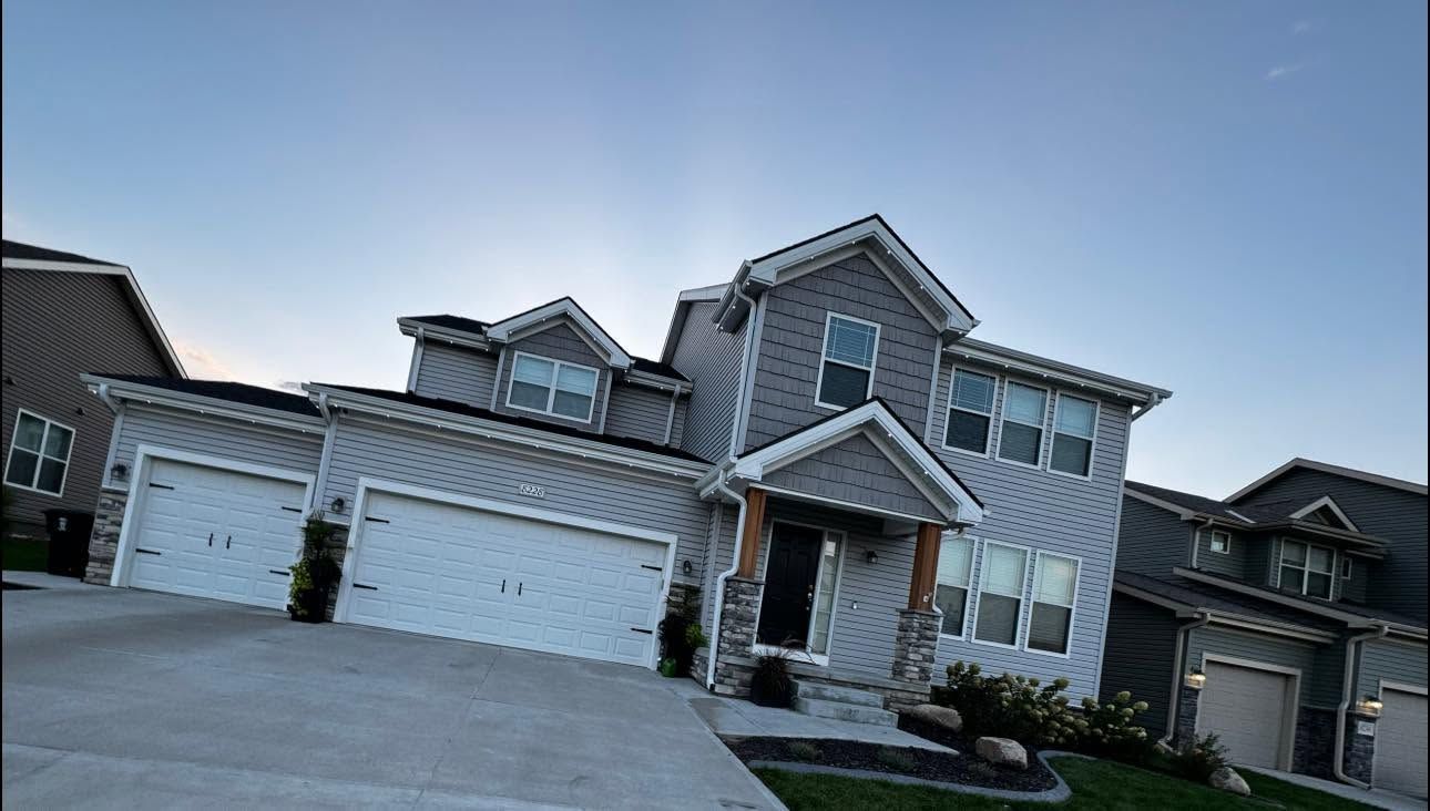 Large gray suburban house with white garage doors and a front porch on a sloped driveway.