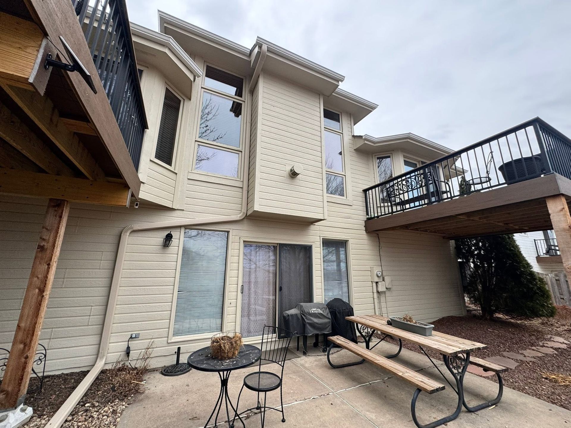 Back patio of a beige two-story house with a table, chairs, and an elevated deck above.
