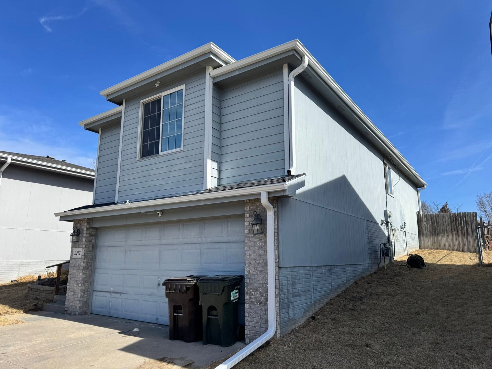 Two-story gray house with a garage, blue sky, and two trash bins in front