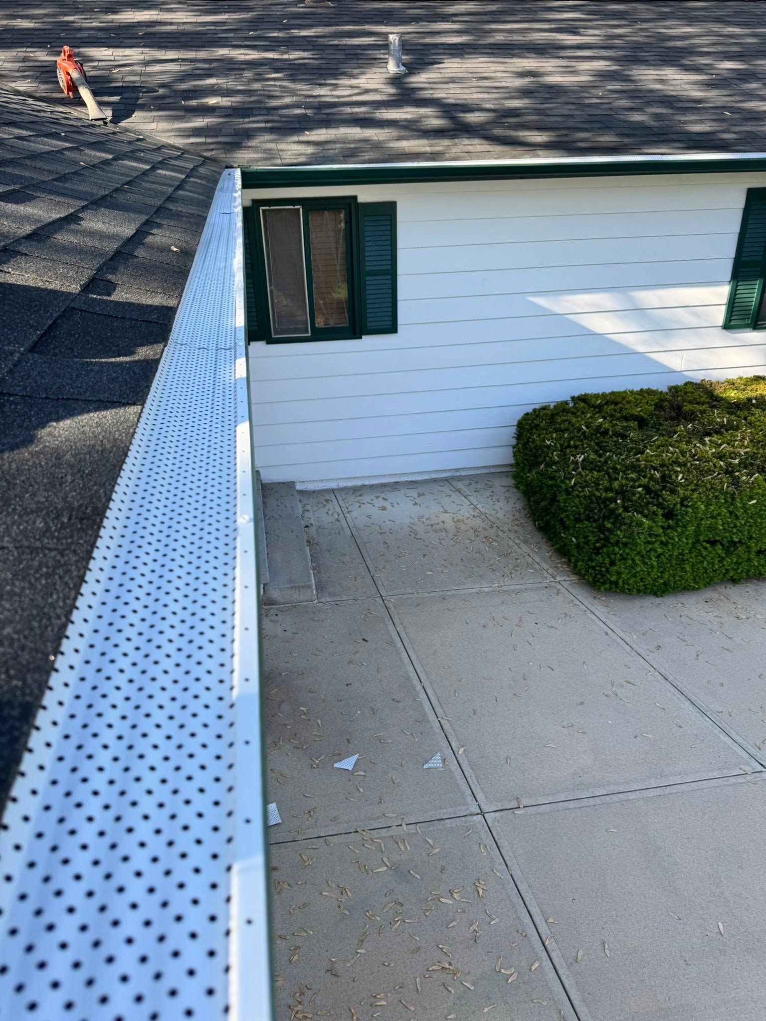 White building with green-trimmed window beside a paved patio and trimmed shrub, seen from above