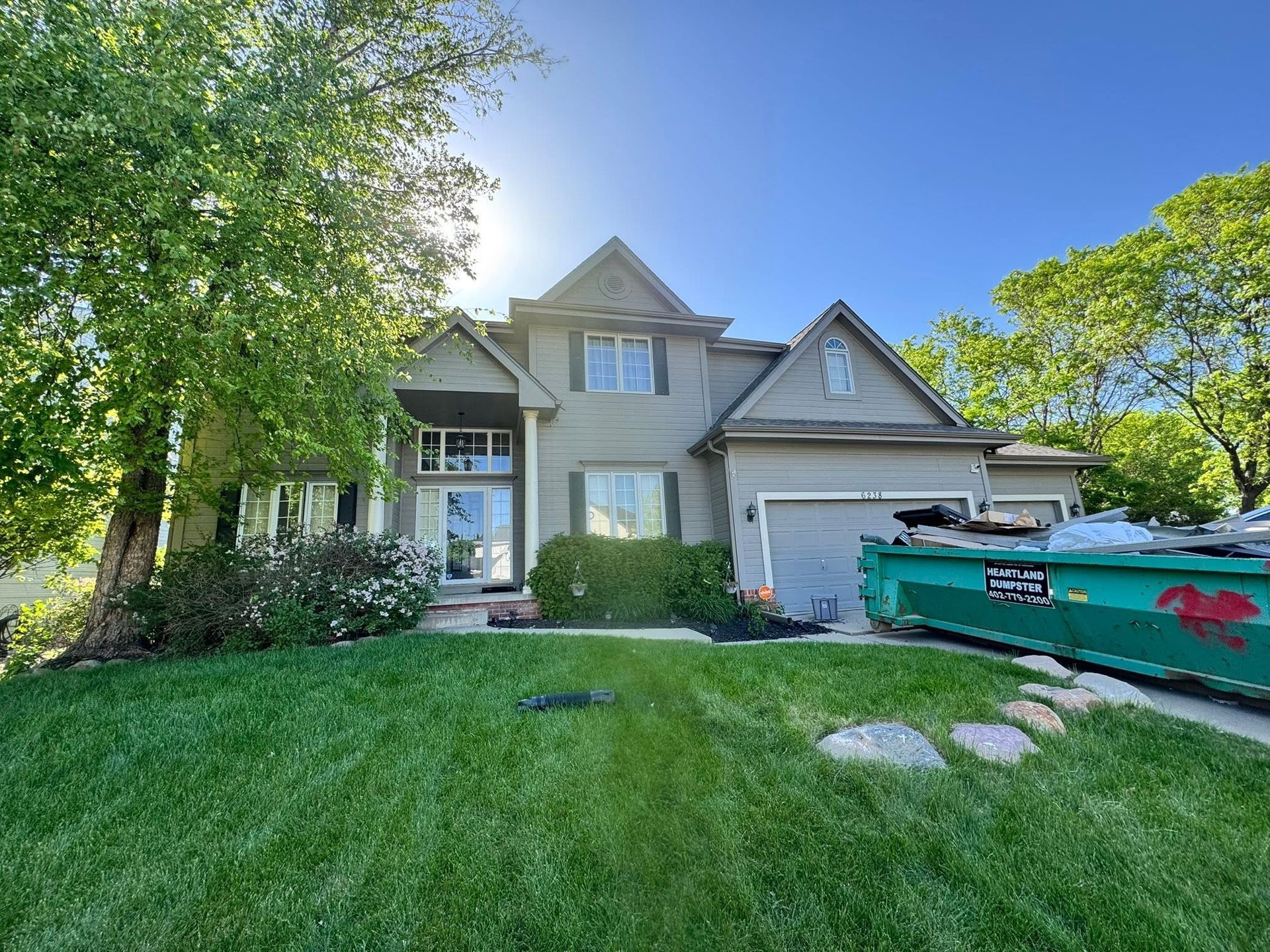 Suburban house with green lawn, trees, and a teal dumpster in the driveway under a blue sky