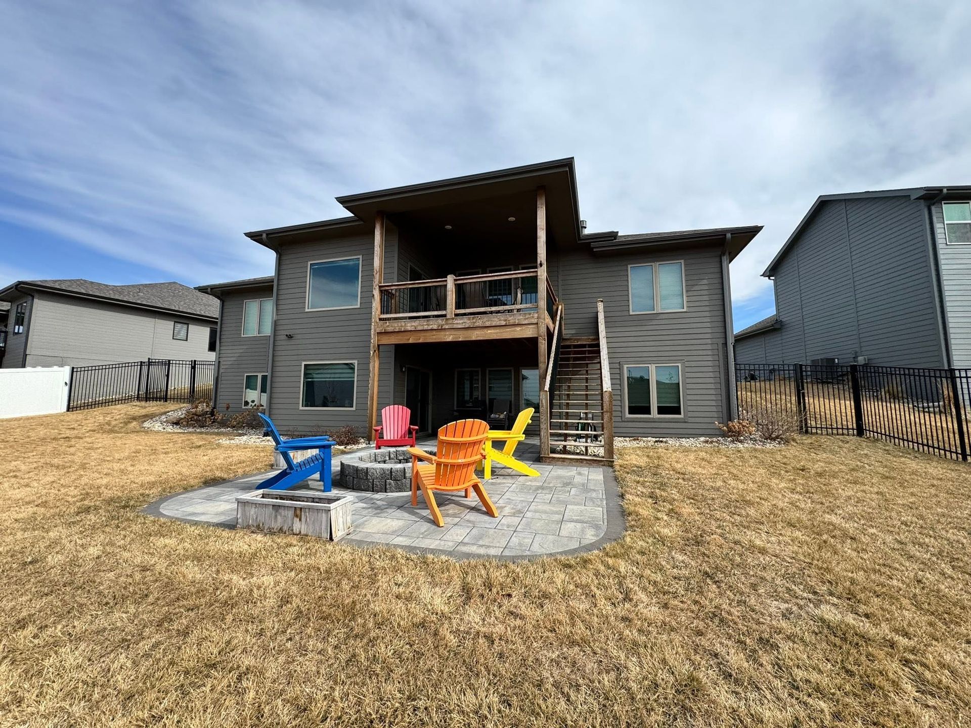 Backyard patio with wooden deck, stone fire pit, and colorful Adirondack chairs beside a house