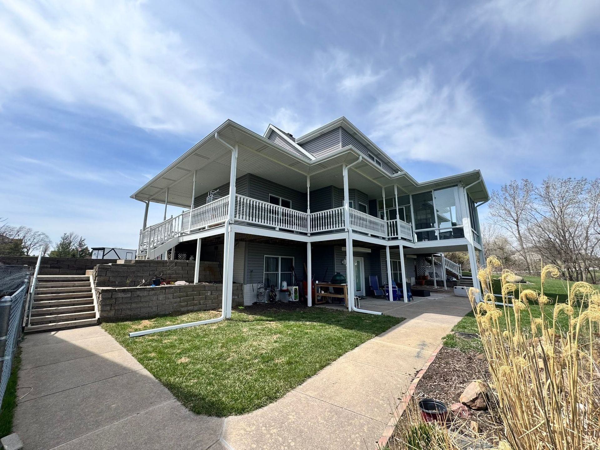 Two-story white house with wraparound porch and stairs, viewed from a landscaped front yard under a blue sky