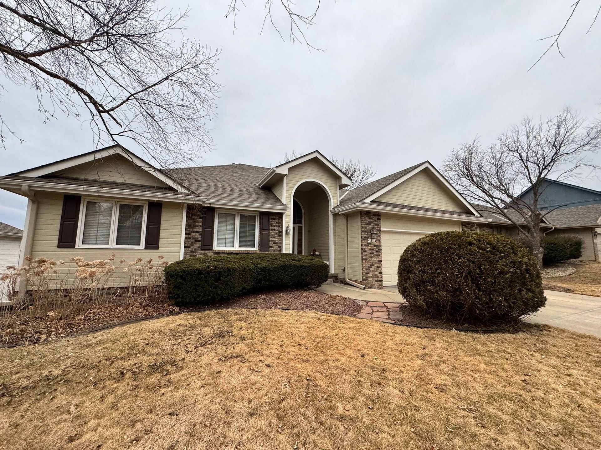 Suburban single-story house with beige siding, brick accents, and a front lawn in winter.