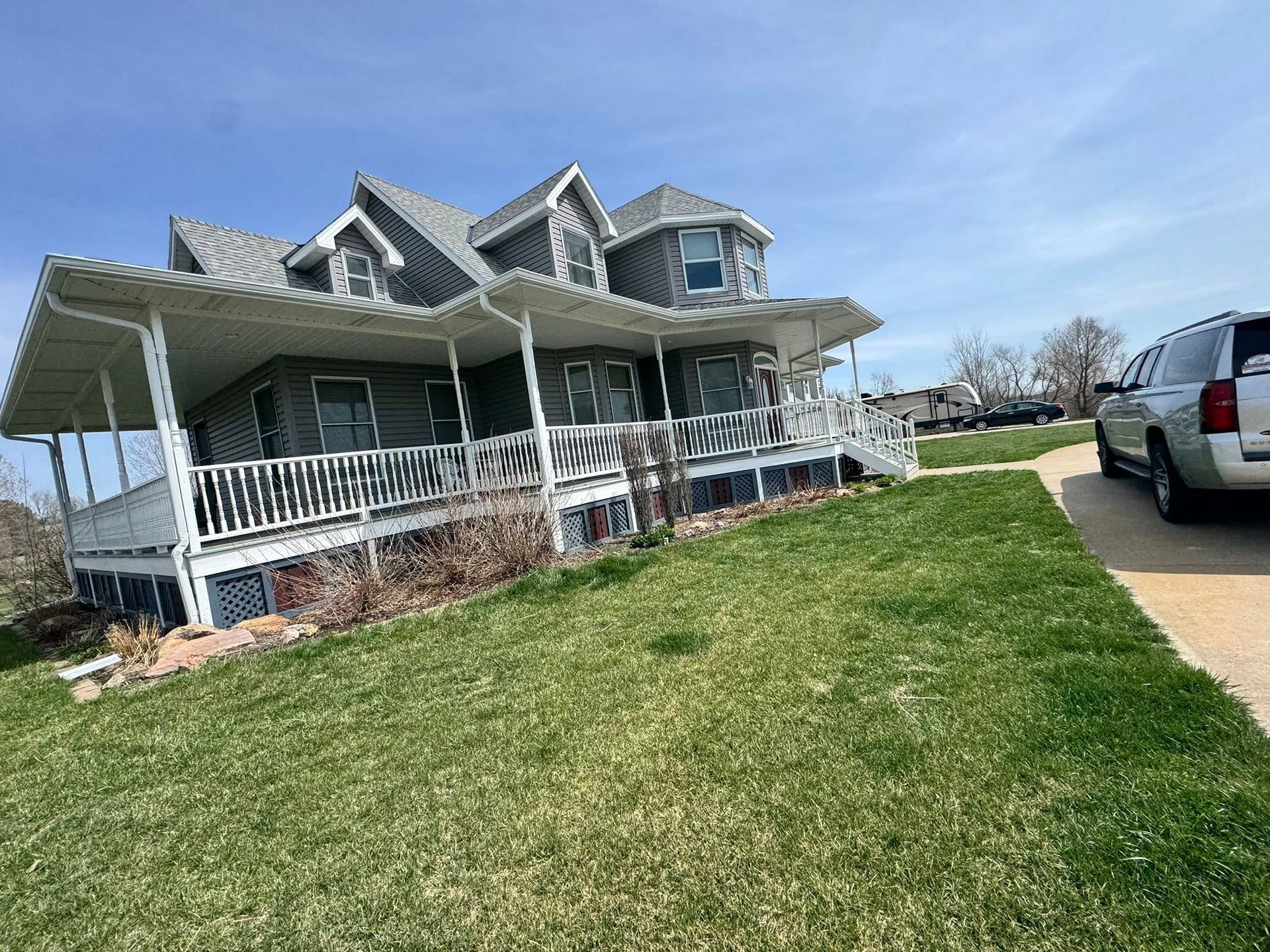 Large gray house with white wraparound porch beside a green lawn and driveway with parked SUV