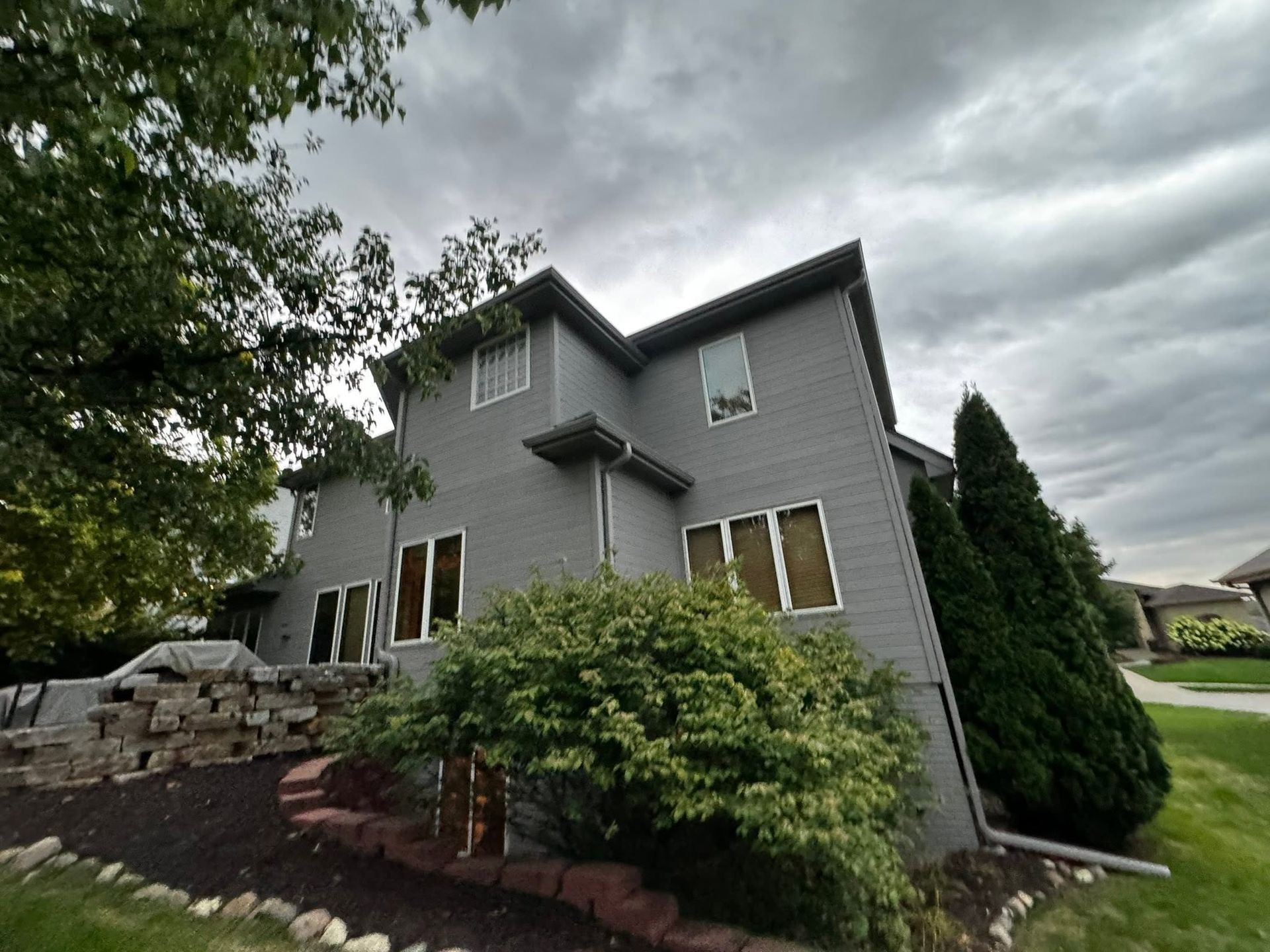 Gray two-story house with stone steps, shrubs, and a cloudy sky in a landscaped yard