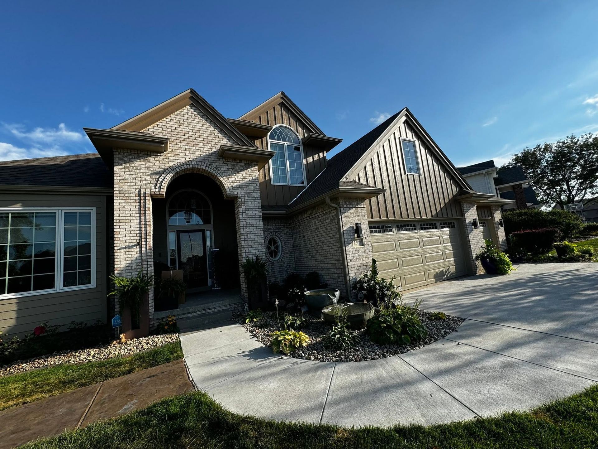 Suburban stone house with arched entry, gabled roof, and curved concrete driveway under a blue sky