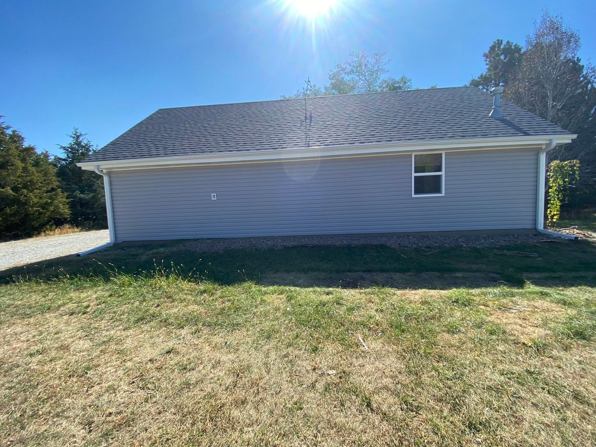 Gray house exterior with a rippled roof, small window, and sunlit yard under a bright blue sky