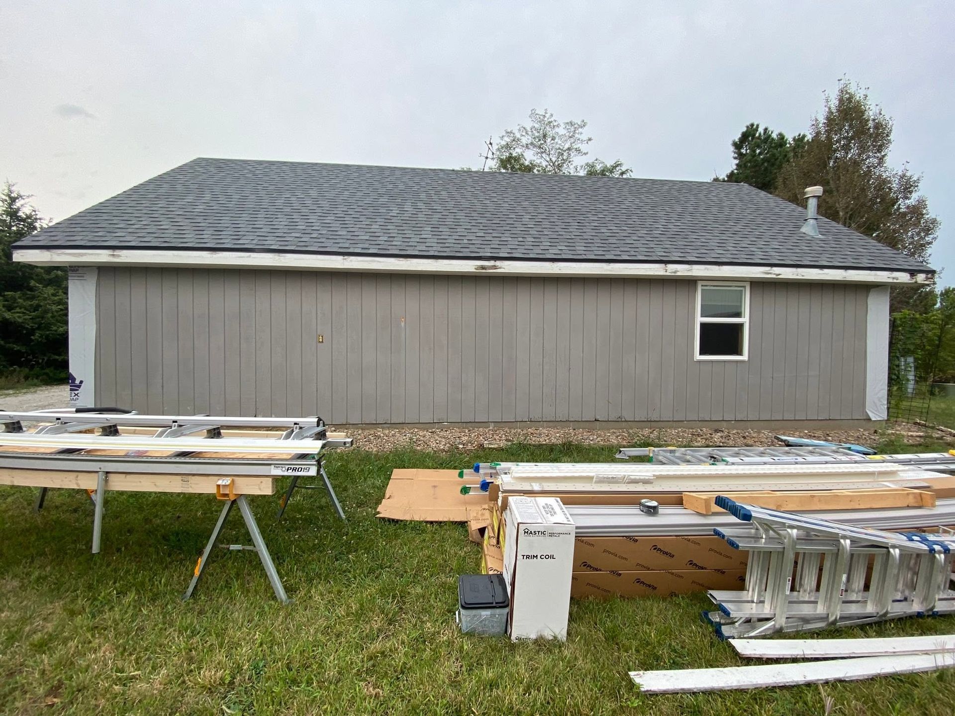 Single-story gray house with a dark roof, viewed from the yard with construction materials in the foreground.