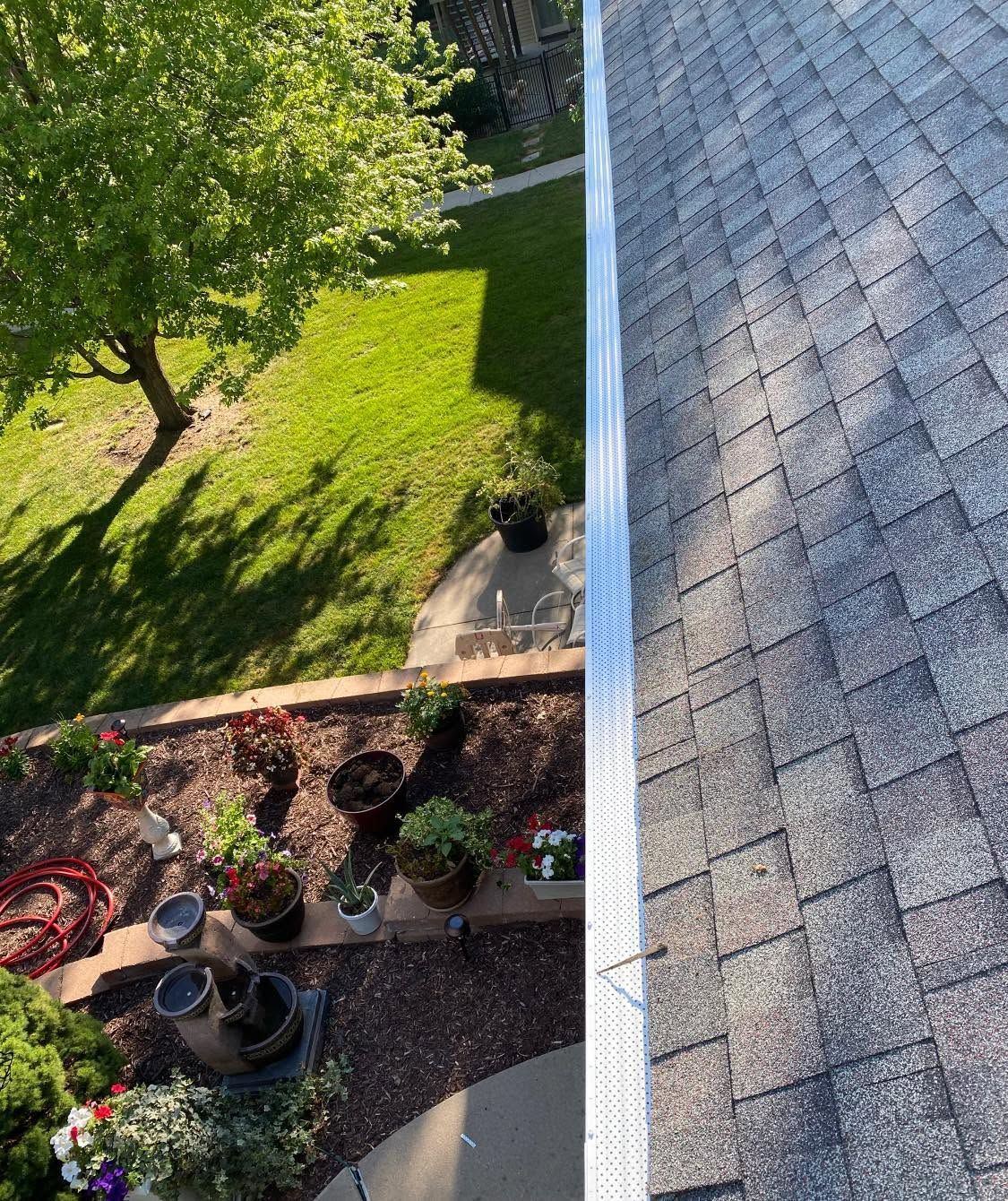 Backyard garden beside a house roof, with green lawn, patio potted plants, and a tree shadow
