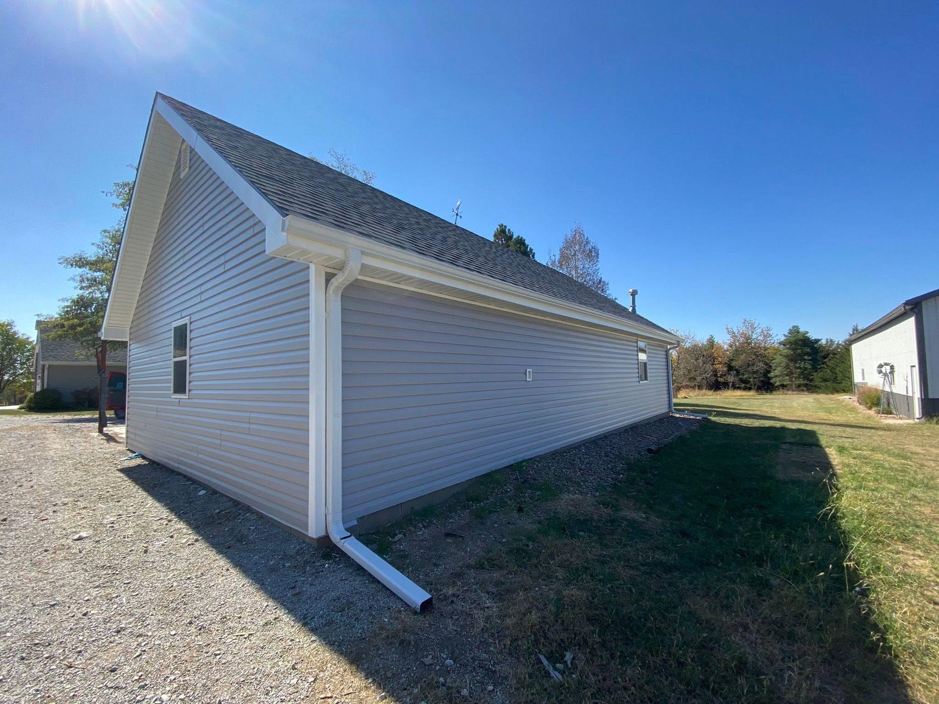 Side view of a white house with a steep roof, gravel drive, and grassy yard under a blue sky