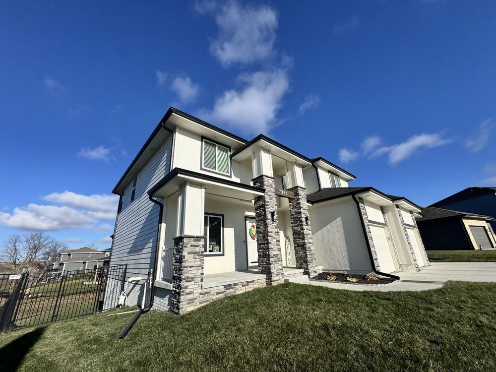 Two-story suburban house with light siding, front porch, and a grassy yard under a blue sky