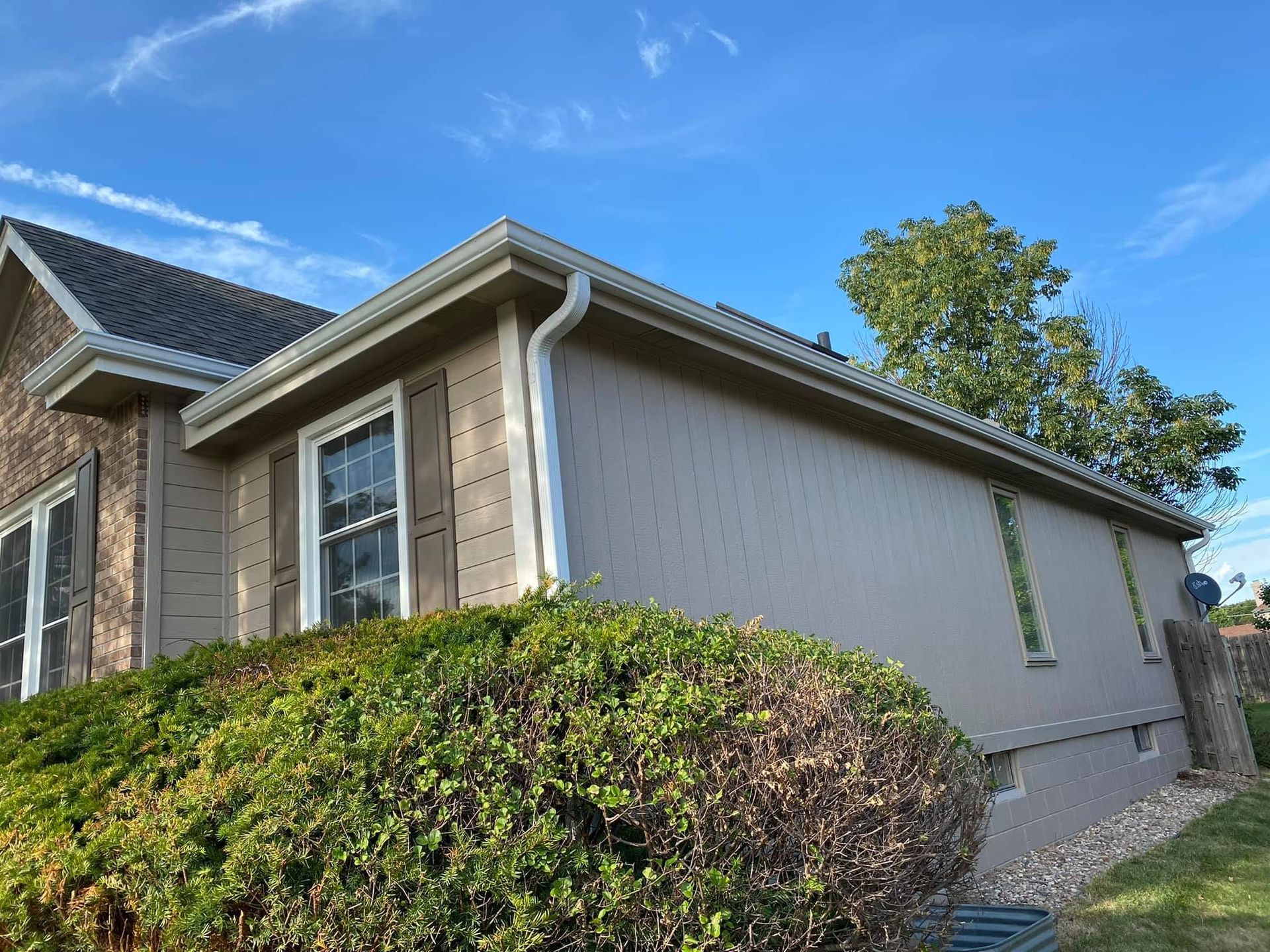 Single-story brick house with gray siding, white trim, and a green hedge under a blue sky