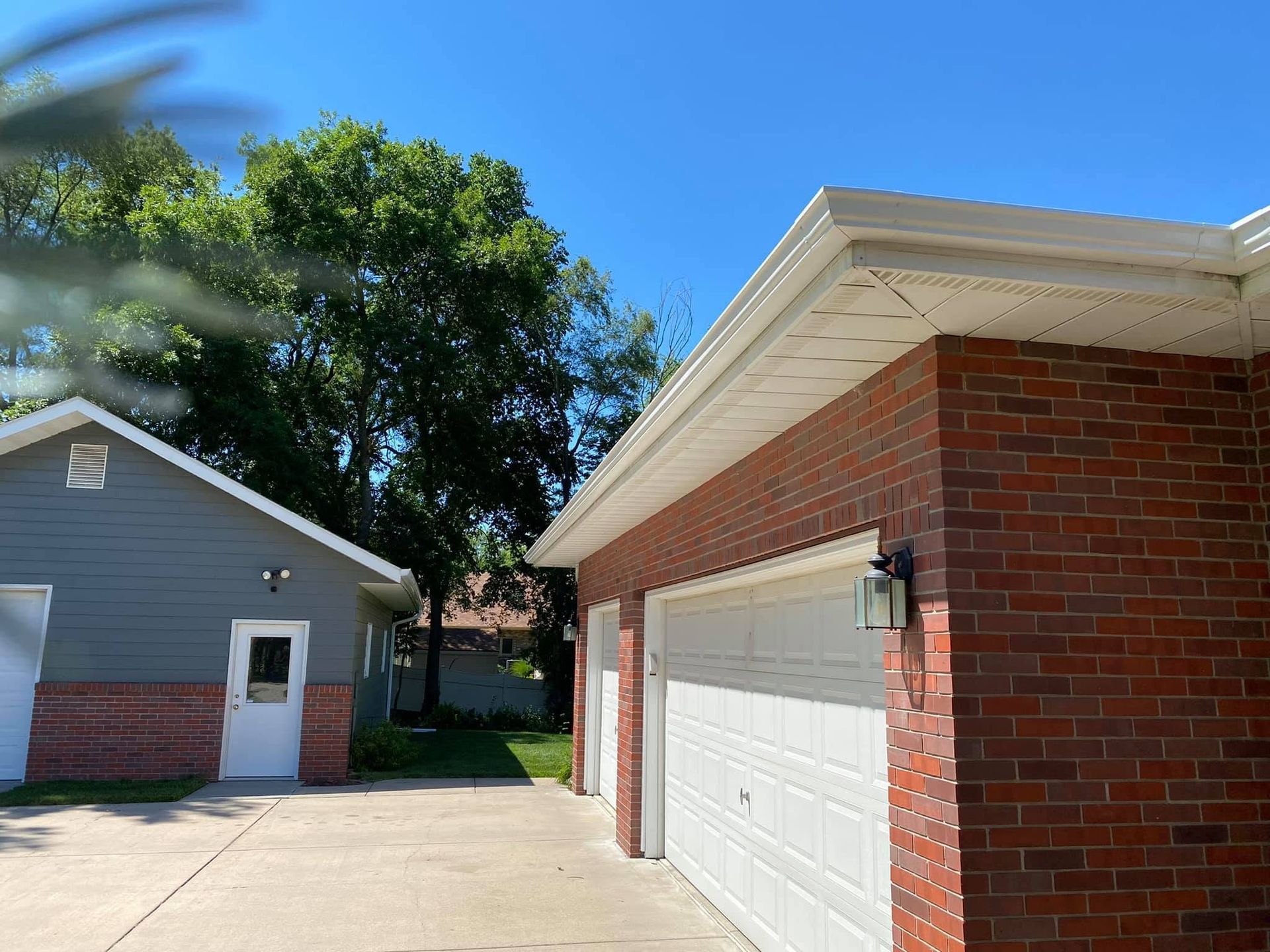 Brick garage beside a gray house on a sunny residential driveway with trees and blue sky