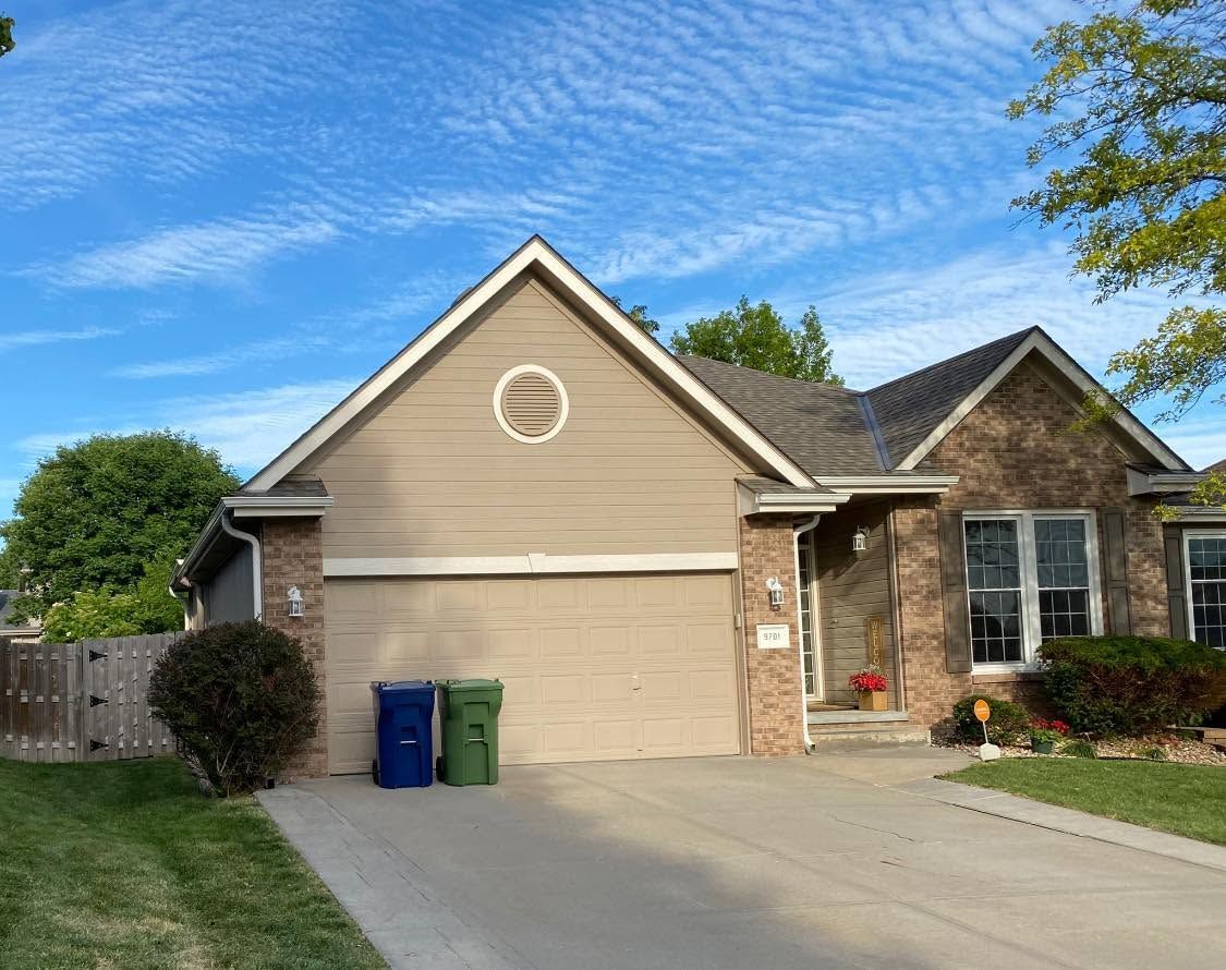 Suburban house with tan garage, driveway, and blue and green bins under a blue sky