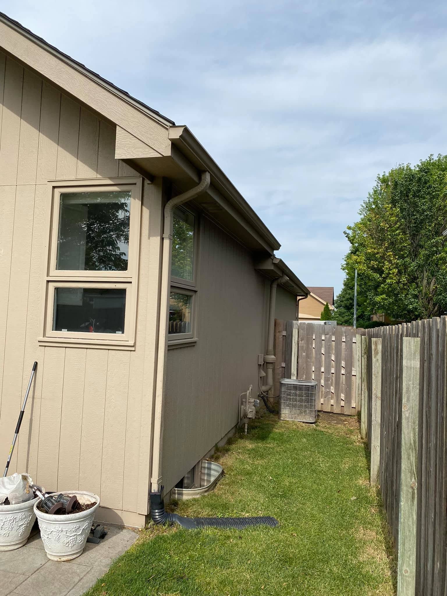 Narrow side yard beside a beige house, with a window, wooden fence, and grass strip under a cloudy sky