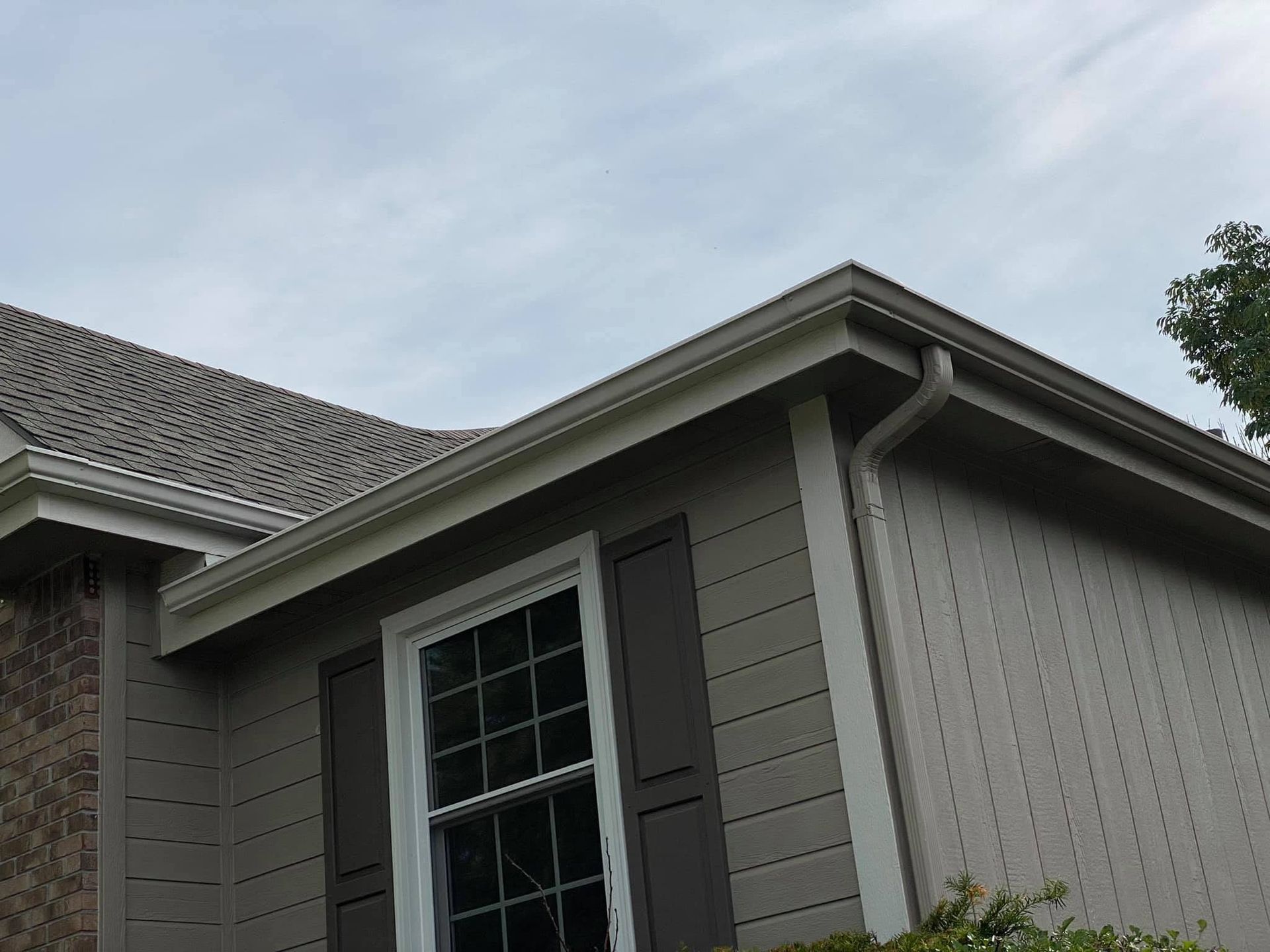 Corner of a gray house with a window, shutters, and roofline against a cloudy sky